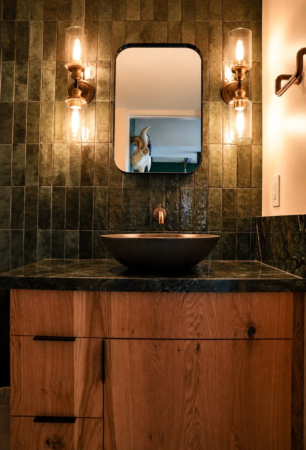 Bathroom vanity with a black vessel sink, wooden cabinet, dark green tiled wall, mirror, and two wall-mounted lights with glass covers.