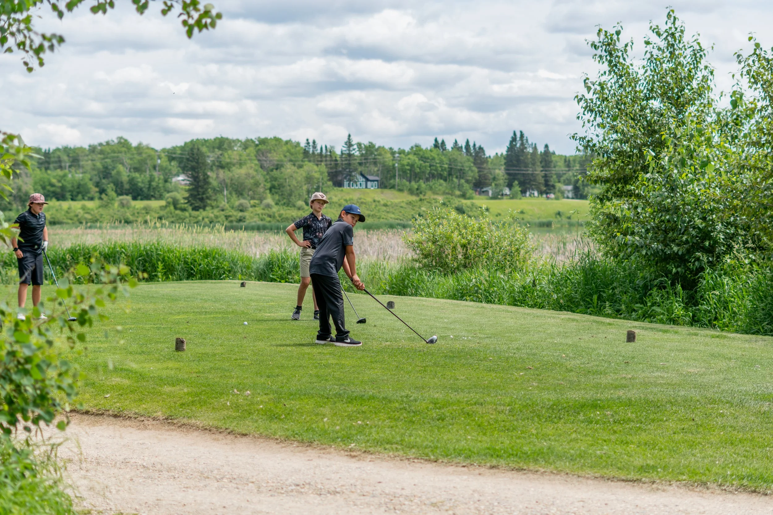Three children playing golf on a green course, surrounded by trees and a cloudy sky.