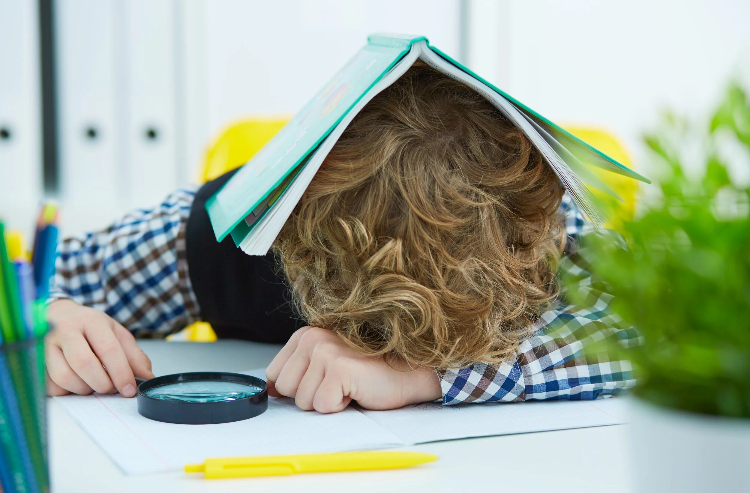 A young girl with curly hair and a checkered shirt has her head down on a desk, with a book balanced on her head. She appears to be studying or concentrating, surrounded by school supplies including yellow pens, markers, and a magnifying glass.
