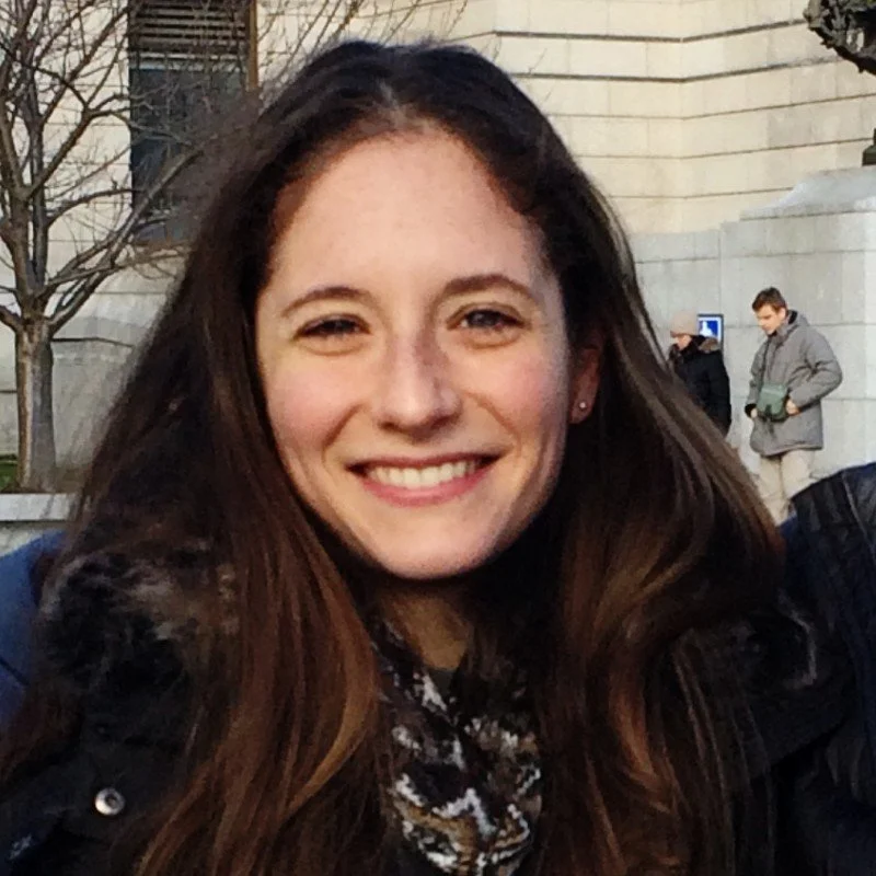 Young woman smiling outdoors with buildings and other people in the background.