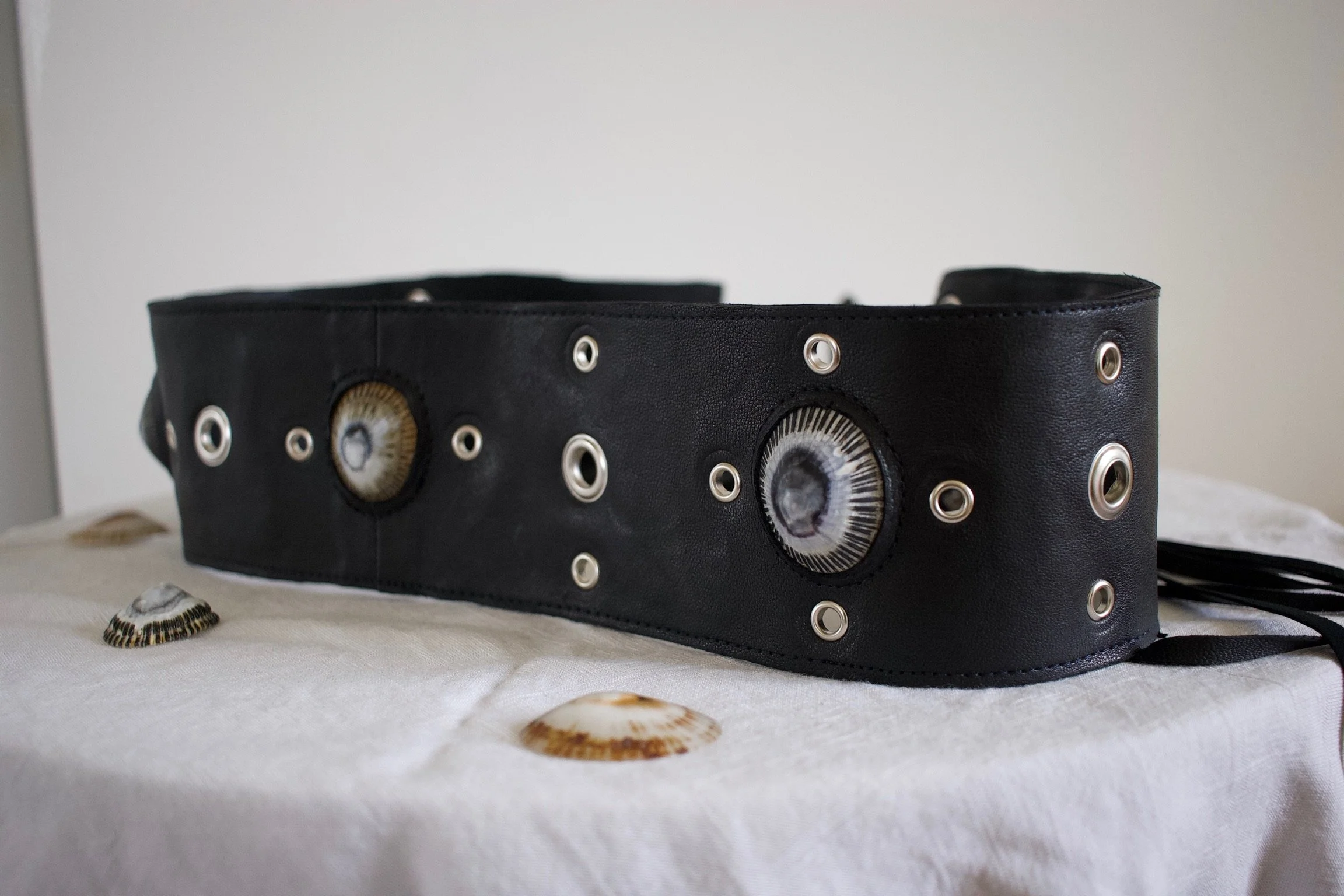 Black belt decorated with seashells and metal eyelets, placed on a white cloth surface with scattered seashells.