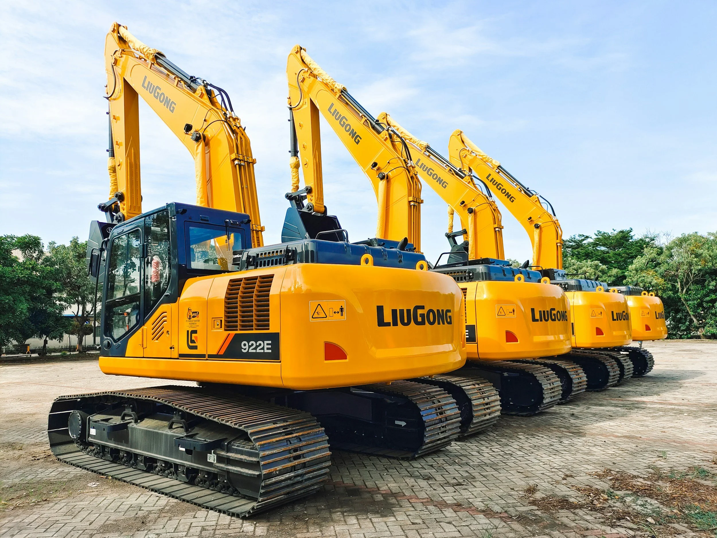 Line of yellow LiuGong excavators parked on paved ground with blue sky and green trees in the background.