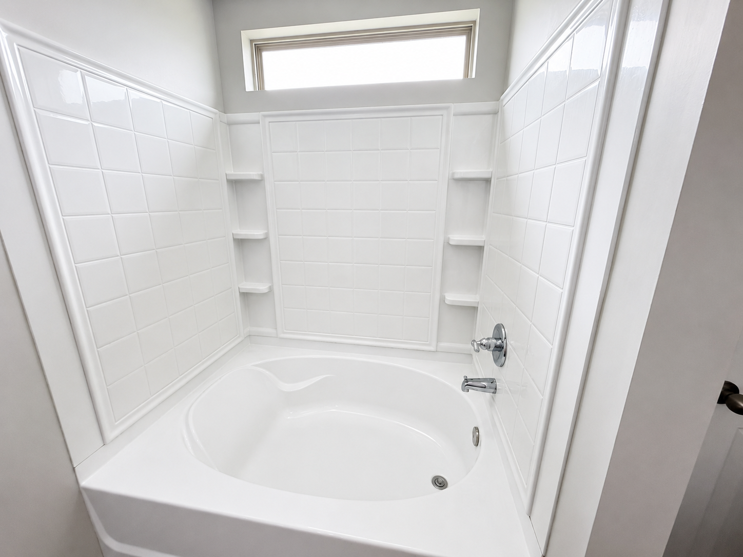 White bathtub with built-in shelving, tiled walls, and a small window above in a bathroom.