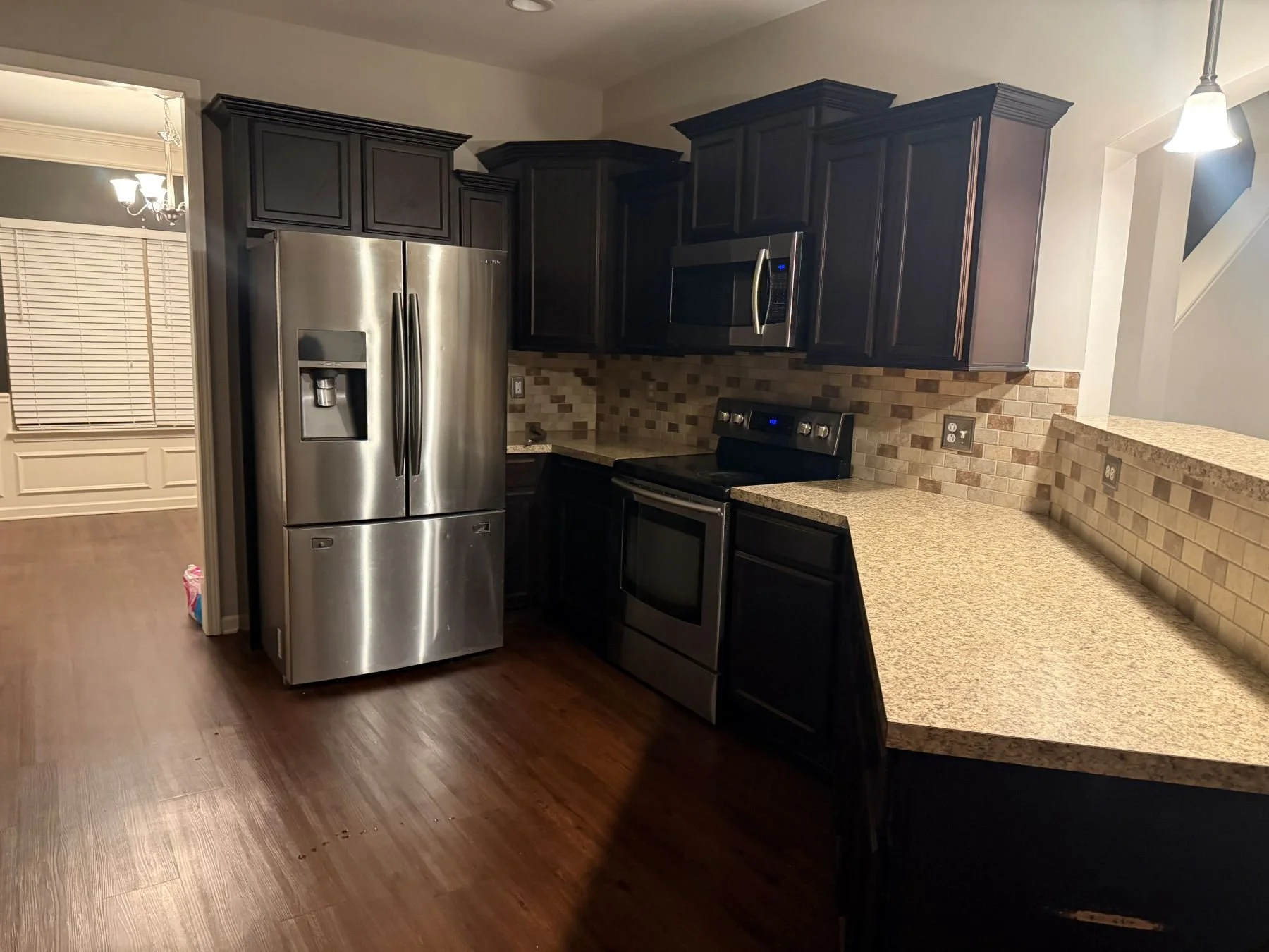 Kitchen with dark cabinets, stainless steel refrigerator and microwave, beige tile backsplash, and granite countertops, hardwood floor, and a small hallway with a dining area and chandelier.
