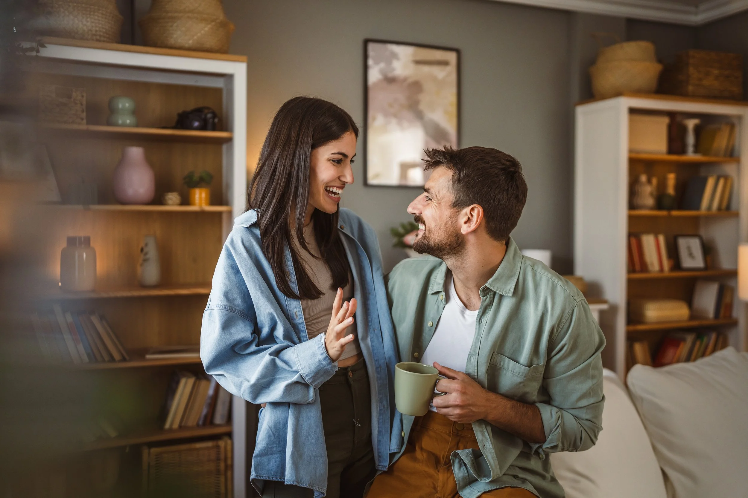 A happy couple sharing a moment in a cozy home, with the woman smiling and talking to the man, who is holding a green mug in front of a bookshelf.