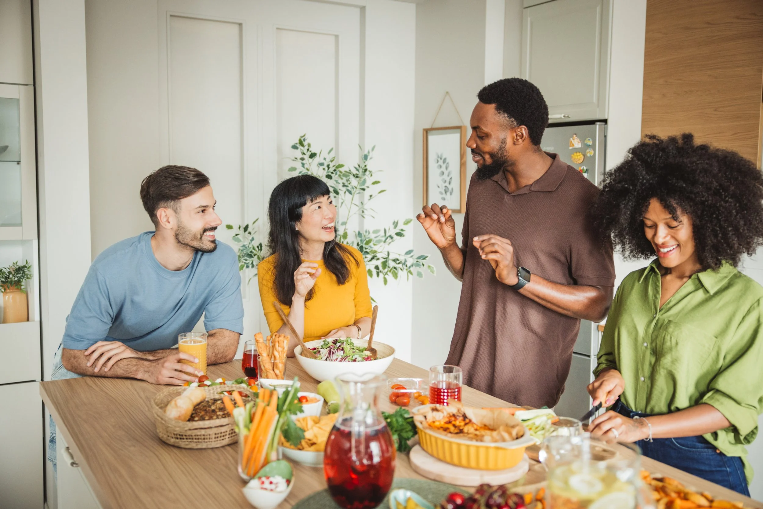 People gathered around a kitchen table enjoying a meal, with food and drinks on the table and smiling faces.