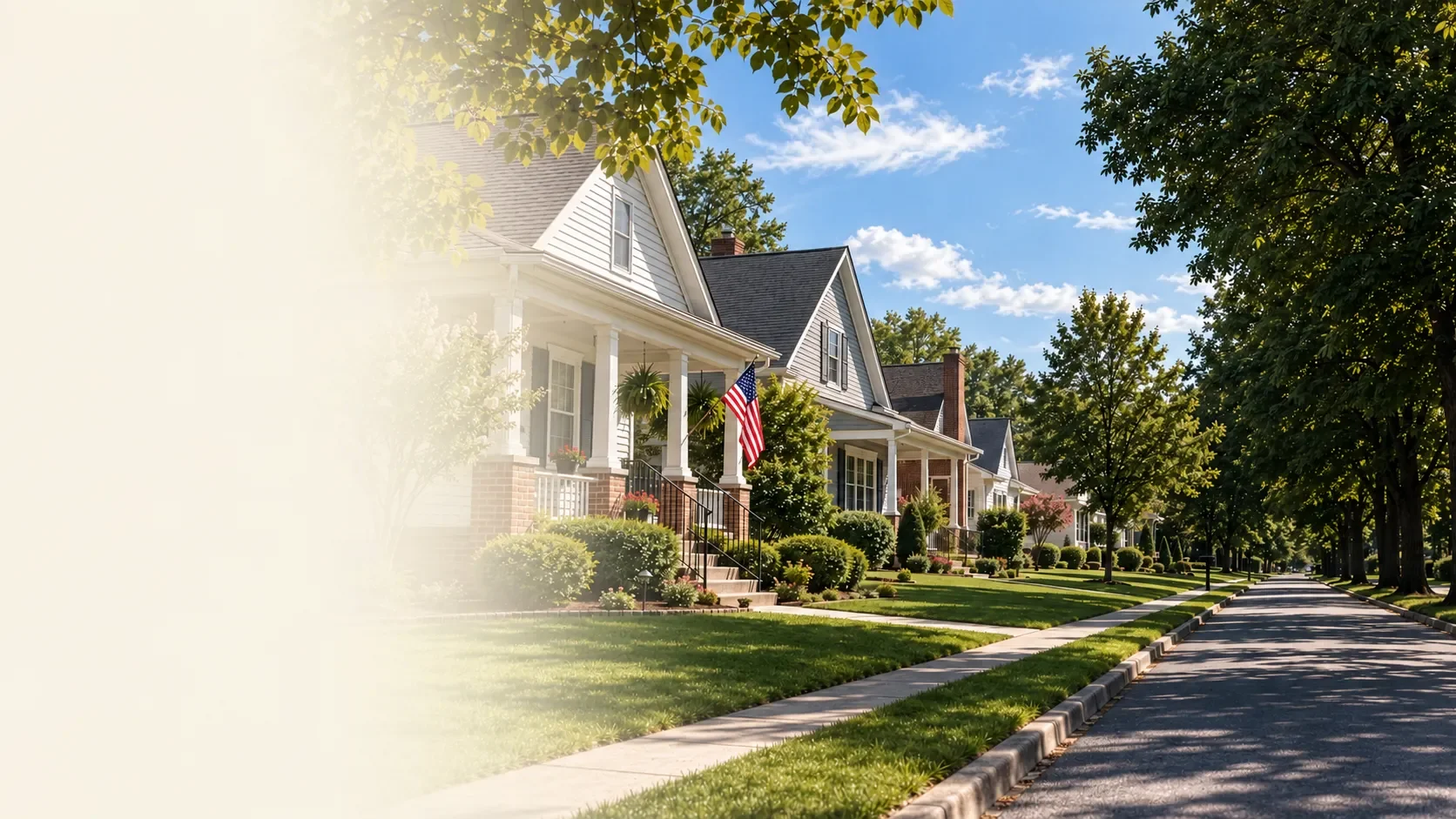 A row of suburban houses with well-maintained lawns, trees, and American flags, lining a quiet residential street on a sunny day with a blue sky and scattered clouds.