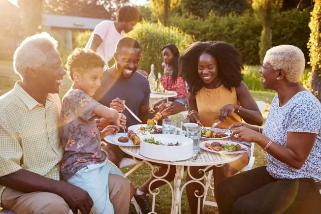 A multigenerational family enjoying a meal outdoors on a sunny day, sitting around a white metal table with food and drinks.