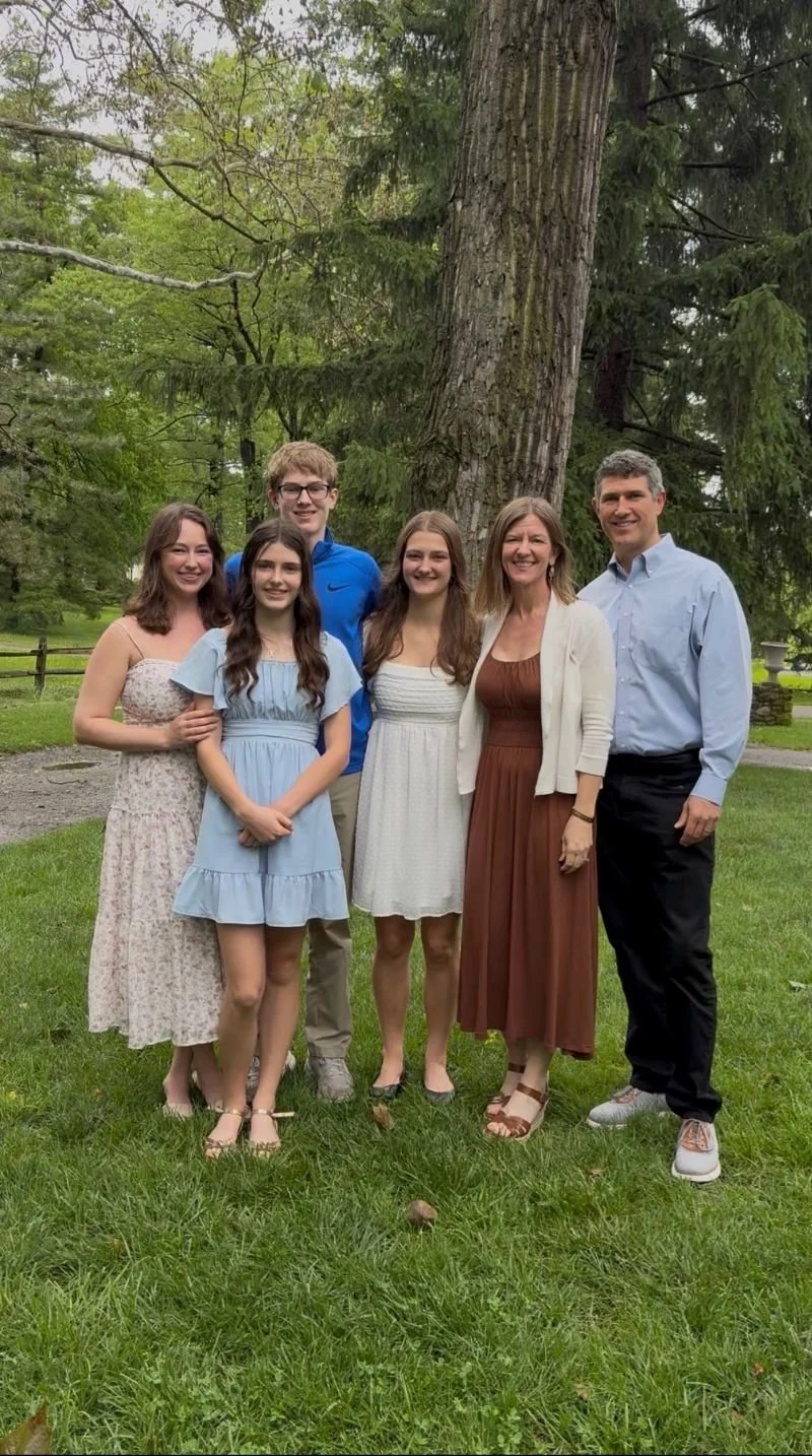 A family of six standing together outdoors in front of a large tree, smiling for a photo. The group includes three young women, two young men, and an older woman, dressed in casual summer clothing.