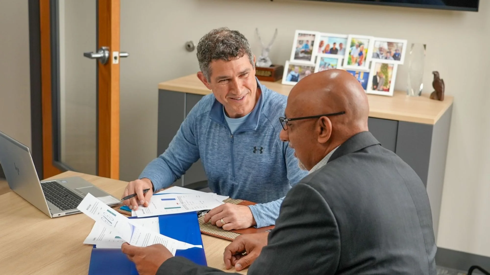 Two men sit at a table with a laptop and paperwork having a conversation about financial planning.