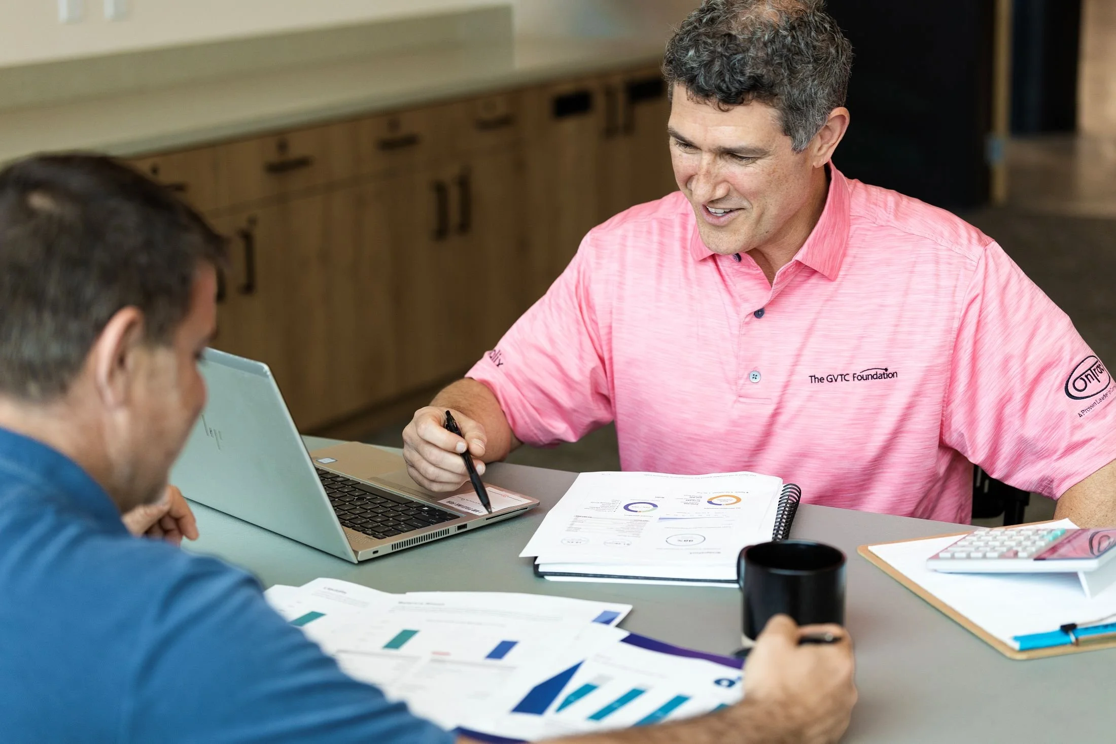 Randy Derenzo in a pink polo shirt and a male client in a blue shirt sitting at a table, engaged in a discussion with documents, a laptop, and charts.