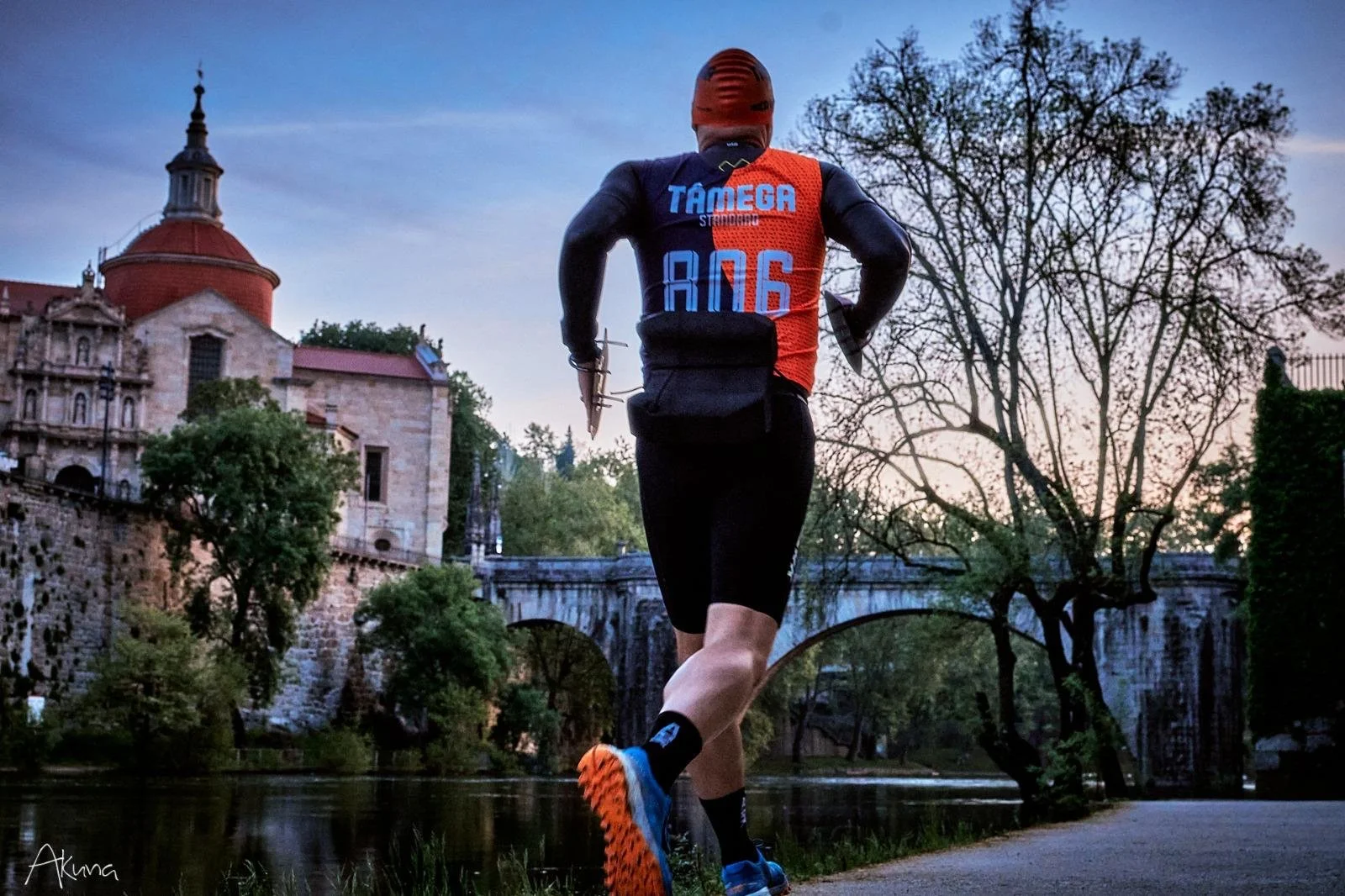 Corredor de costas, com uniforme de corrida, usando capacete, correndo ao lado de rio e árvores ao entardecer, fundo com ponte antiga e edifícios históricos.