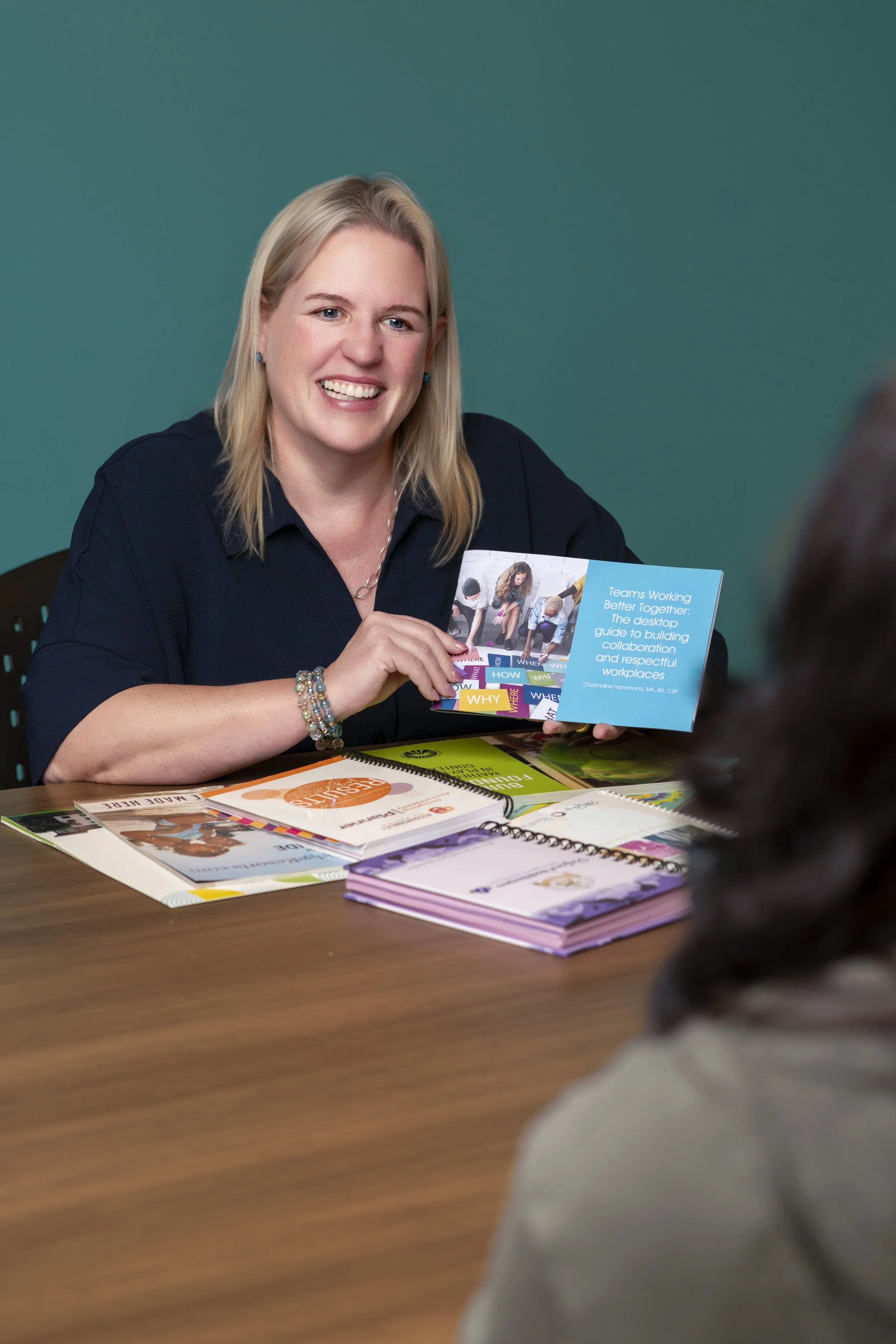 A woman with blonde hair smiling and talking to someone across a table, holding a pamphlet titled 'Teams Working Better Together: The desktop guide to building collaboration and respectful workplaces.' The table has various pamphlets and notebooks on it.