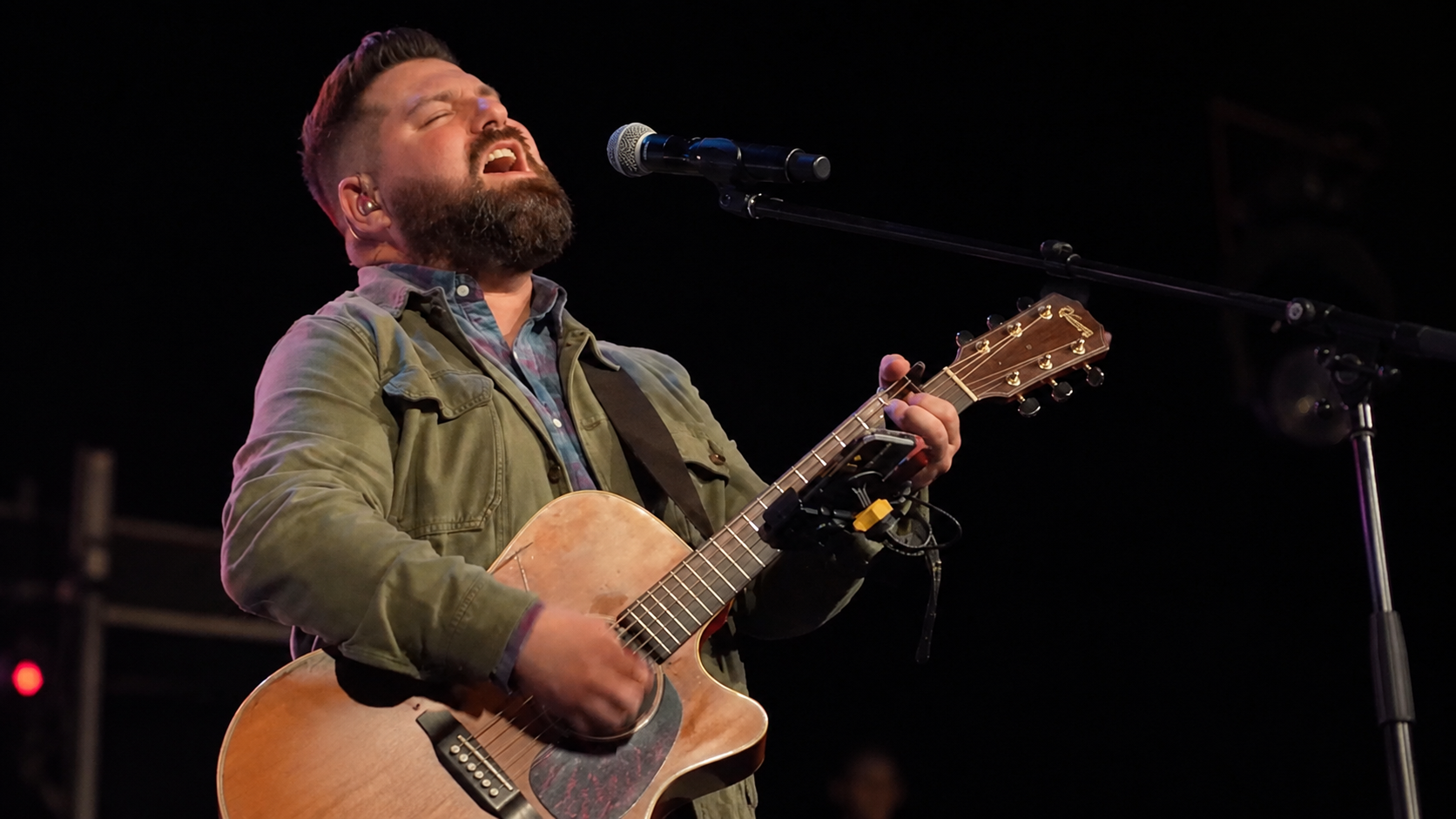 A man with a beard is performing on stage, singing with eyes closed while playing an acoustic guitar. He is wearing a greenish jacket over a plaid shirt near a microphone.