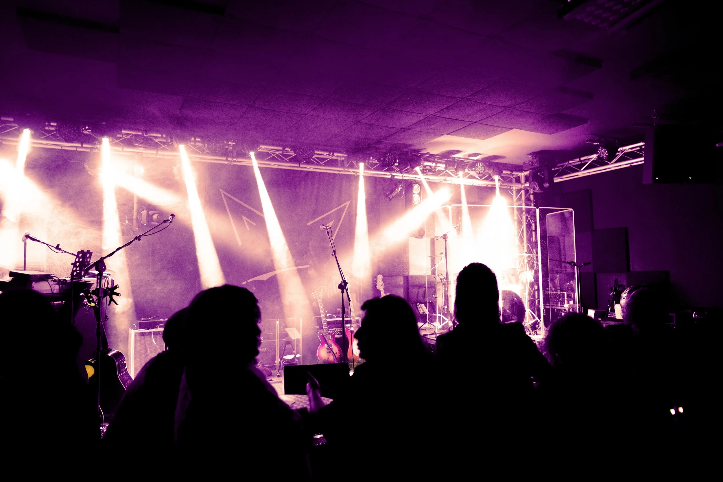 Concert stage with bright lights, musical instruments, and silhouettes of audience members.