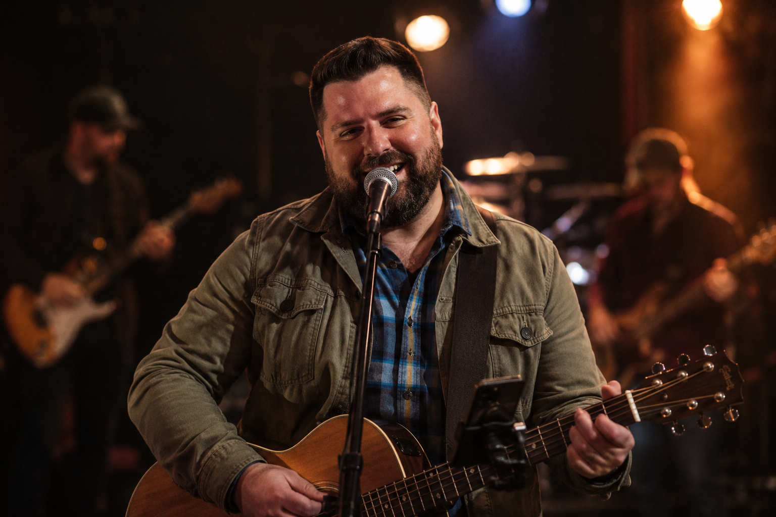 A man with a beard and short dark hair singing into a microphone while playing an acoustic guitar on stage. Two other musicians are blurred in the background, playing electric guitars.