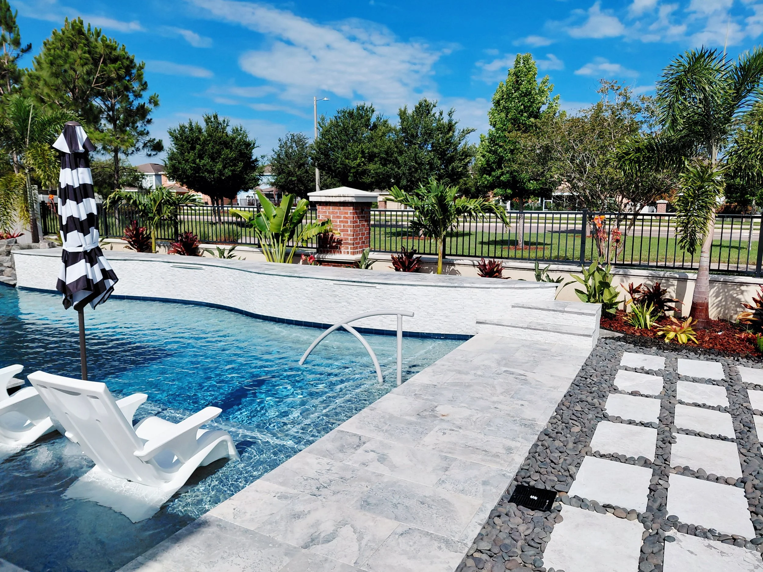 Riverview, FL-Custom travertine pool deck and pathway installation with integrated drainage. Canopy palms and ornamental plantings frame the space, creating depth and a resort-style atmosphere.