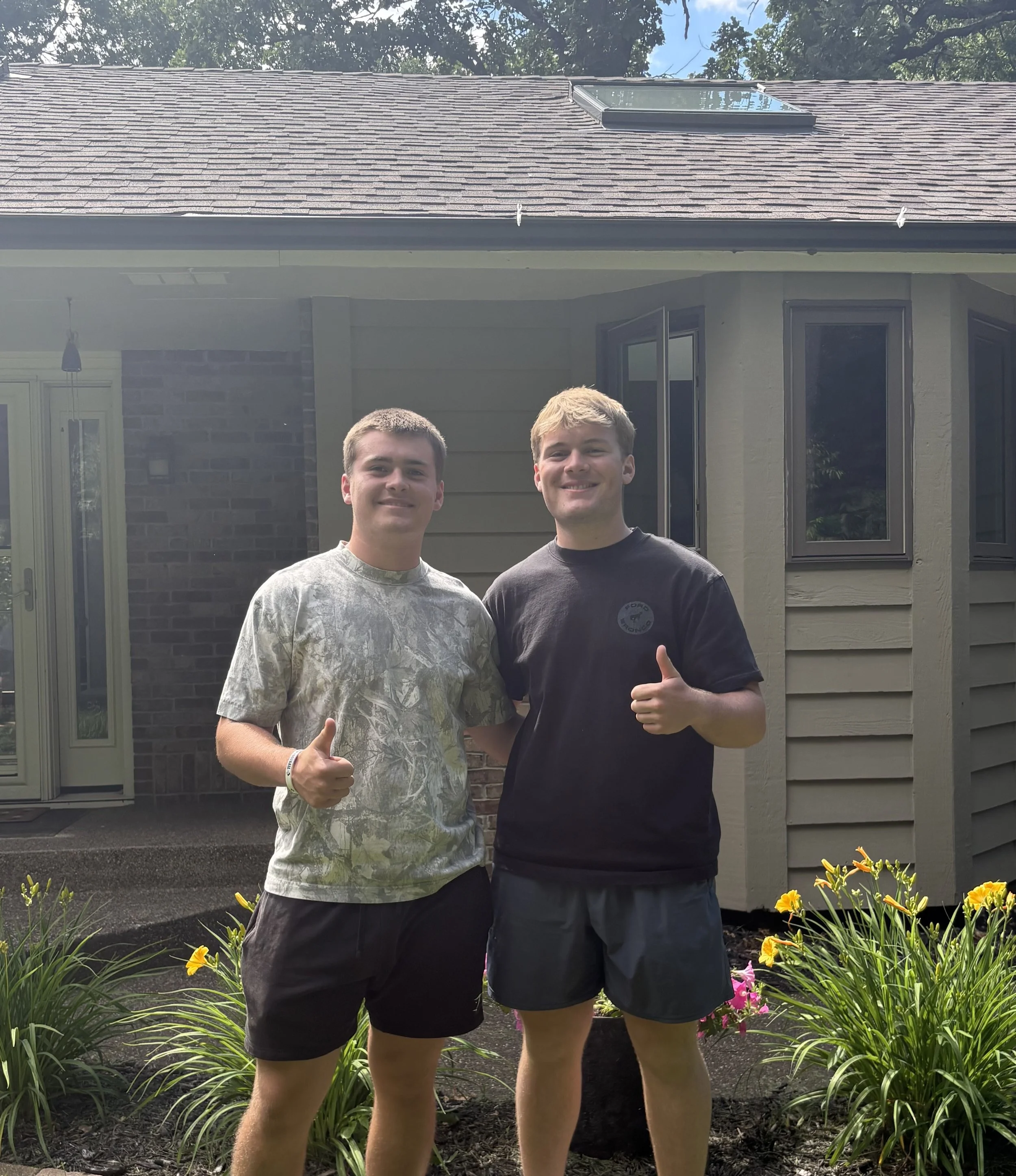 Two young men standing outside in front of a house, smiling and giving thumbs-up, with colorful flowers and green plants in the garden.