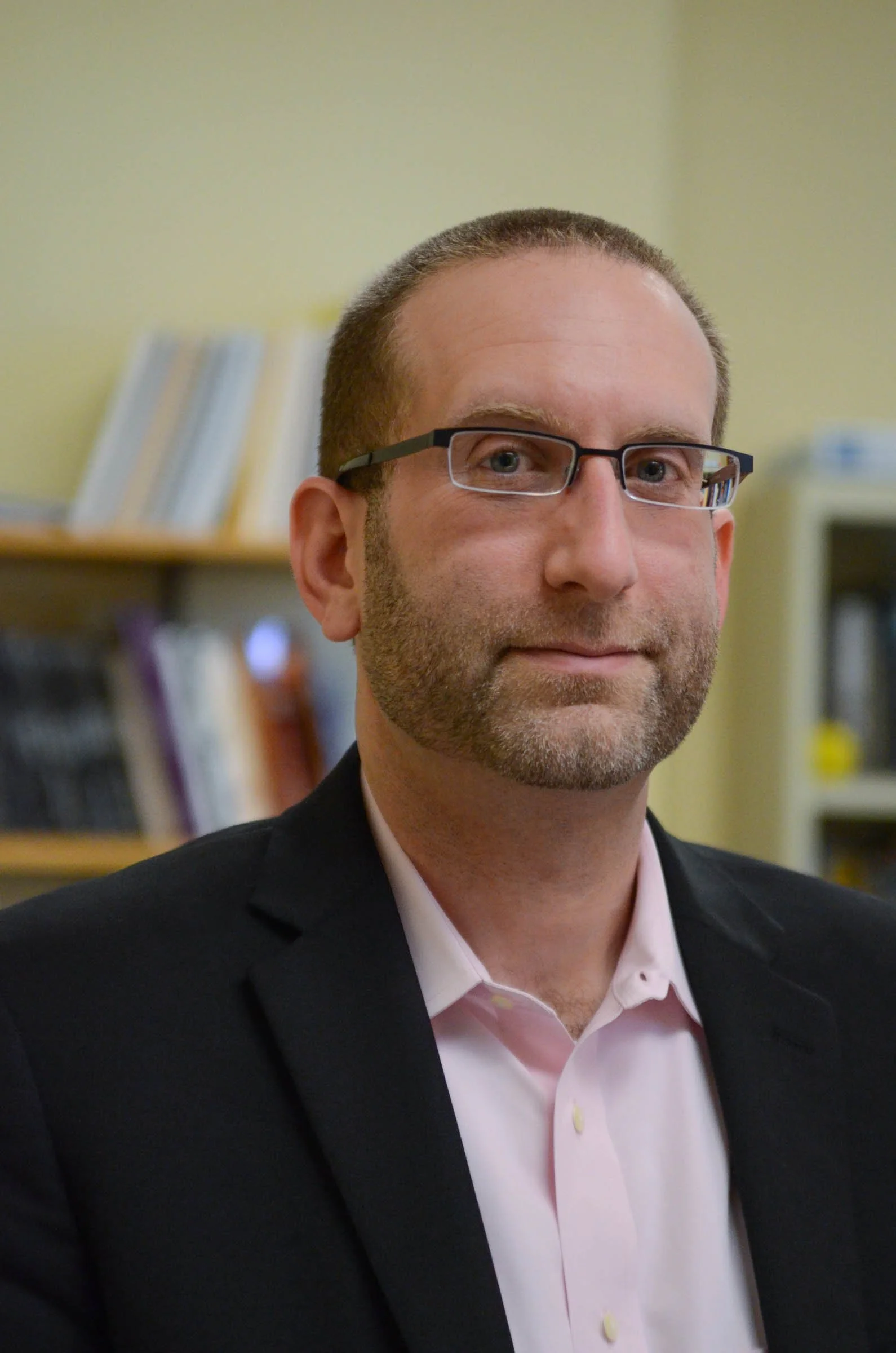 Close-up of a man wearing glasses, a pink dress shirt, and a dark suit jacket, with a blurred bookshelf in the background.