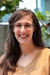 A woman with long brown hair, wearing glasses and earrings, smiling outdoors with greenery and a building in the background.