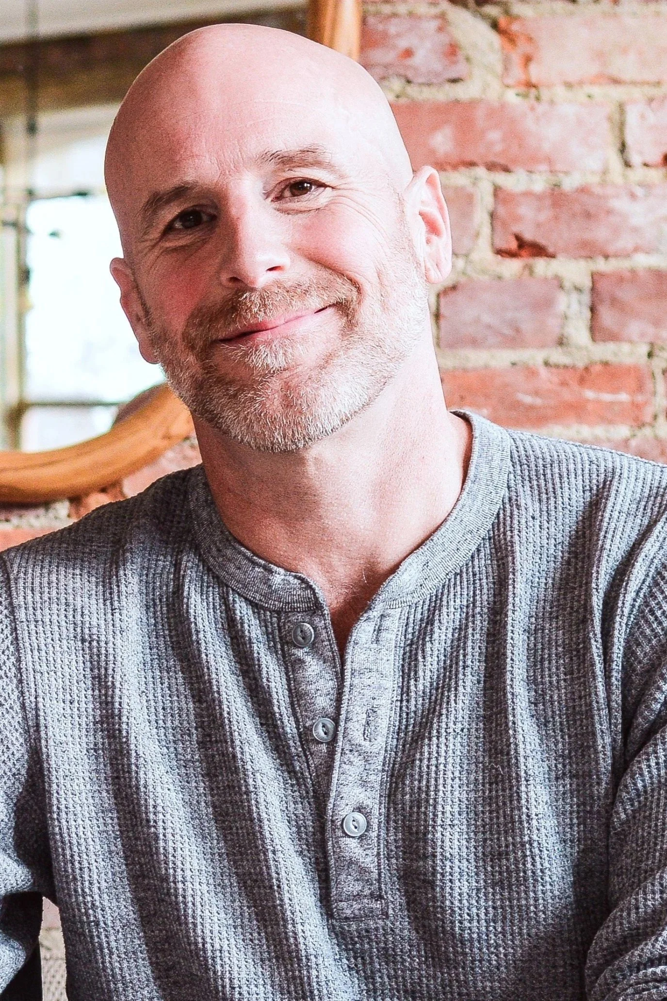 A smiling middle-aged man with a bald head, a beard, and wearing a gray, button-up shirt standing in front of a brick wall.