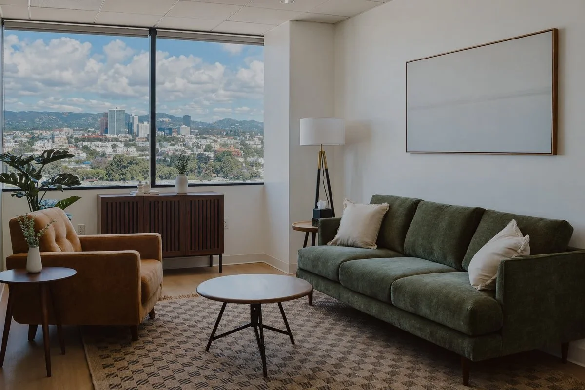 Modern living room with a large window showcasing a cityscape, featuring a green sofa, a tan armchair, a wooden coffee table, a side table with a small vase, and a floor lamp, with abstract artwork on the wall.