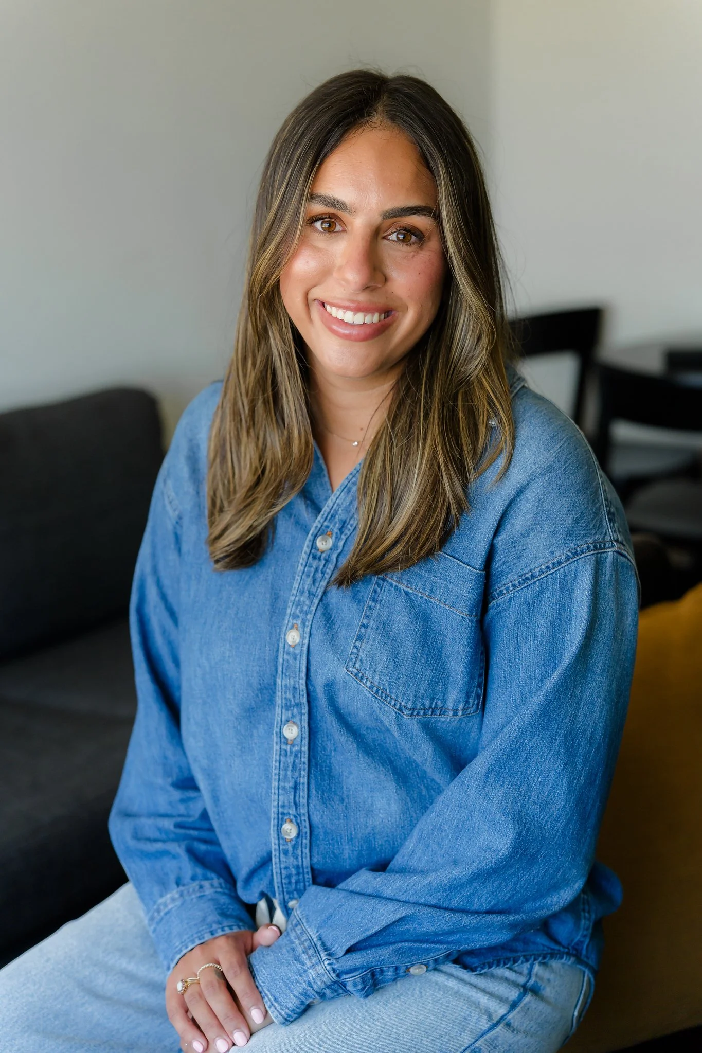 A woman with long brown hair wearing a blue denim button-up shirt, sitting on a dark couch, smiling at the camera with a neutral background.