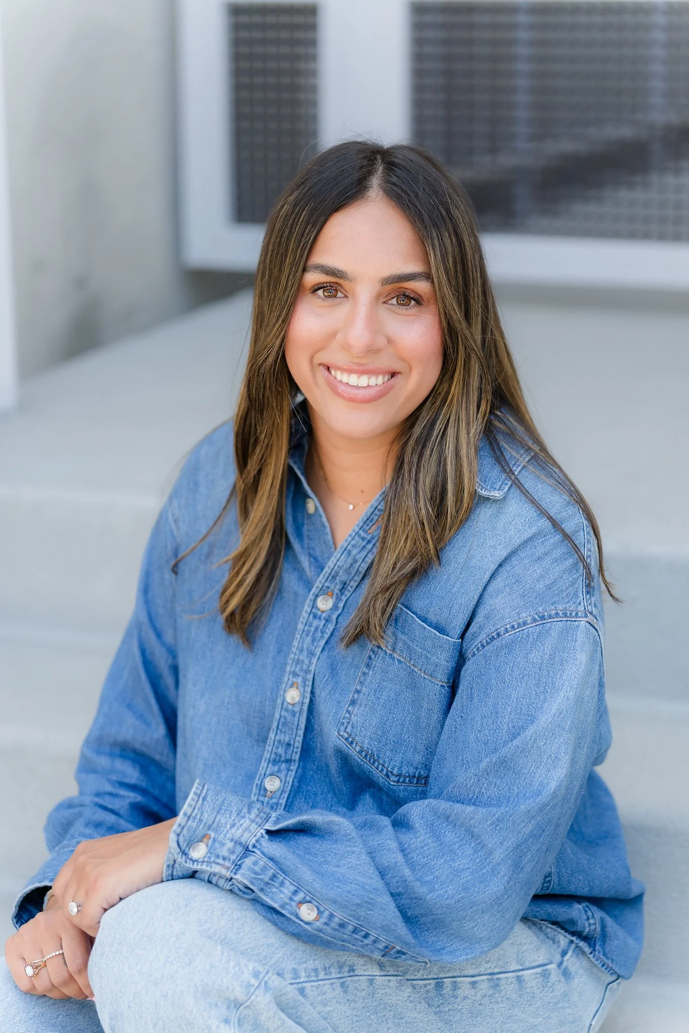 A young woman with long brown hair and brown eyes, smiling, wearing a denim shirt and light blue jeans, sitting outdoors with a modern building and window in the background.