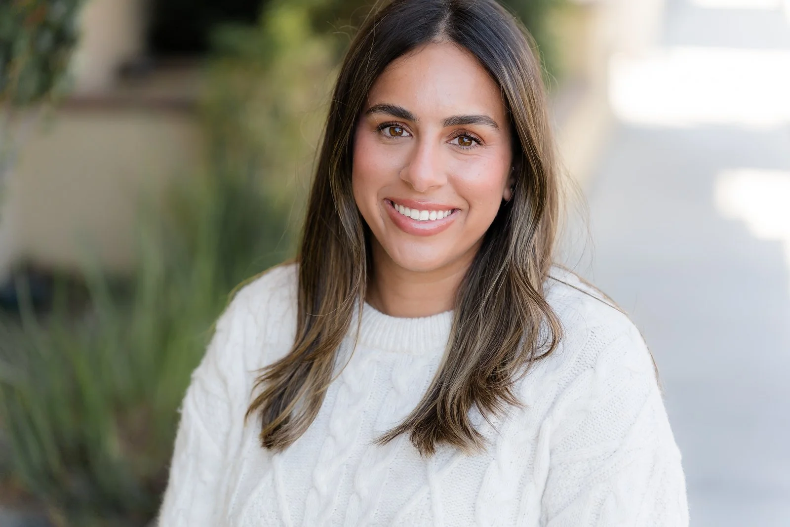 A young woman with long brown hair, brown eyes, and a warm smile, wearing a white cable-knit sweater, outdoors with blurred greenery and pathway in background.
