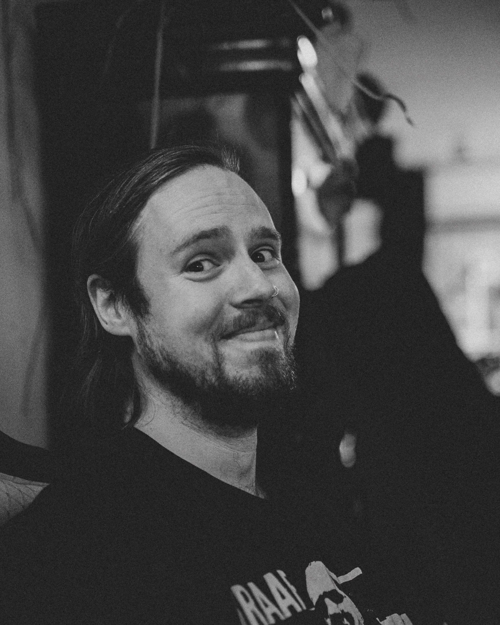 Black and white photo of a smiling man with a beard and nose ring, looking upward, with a blurred background that includes a person and some objects.