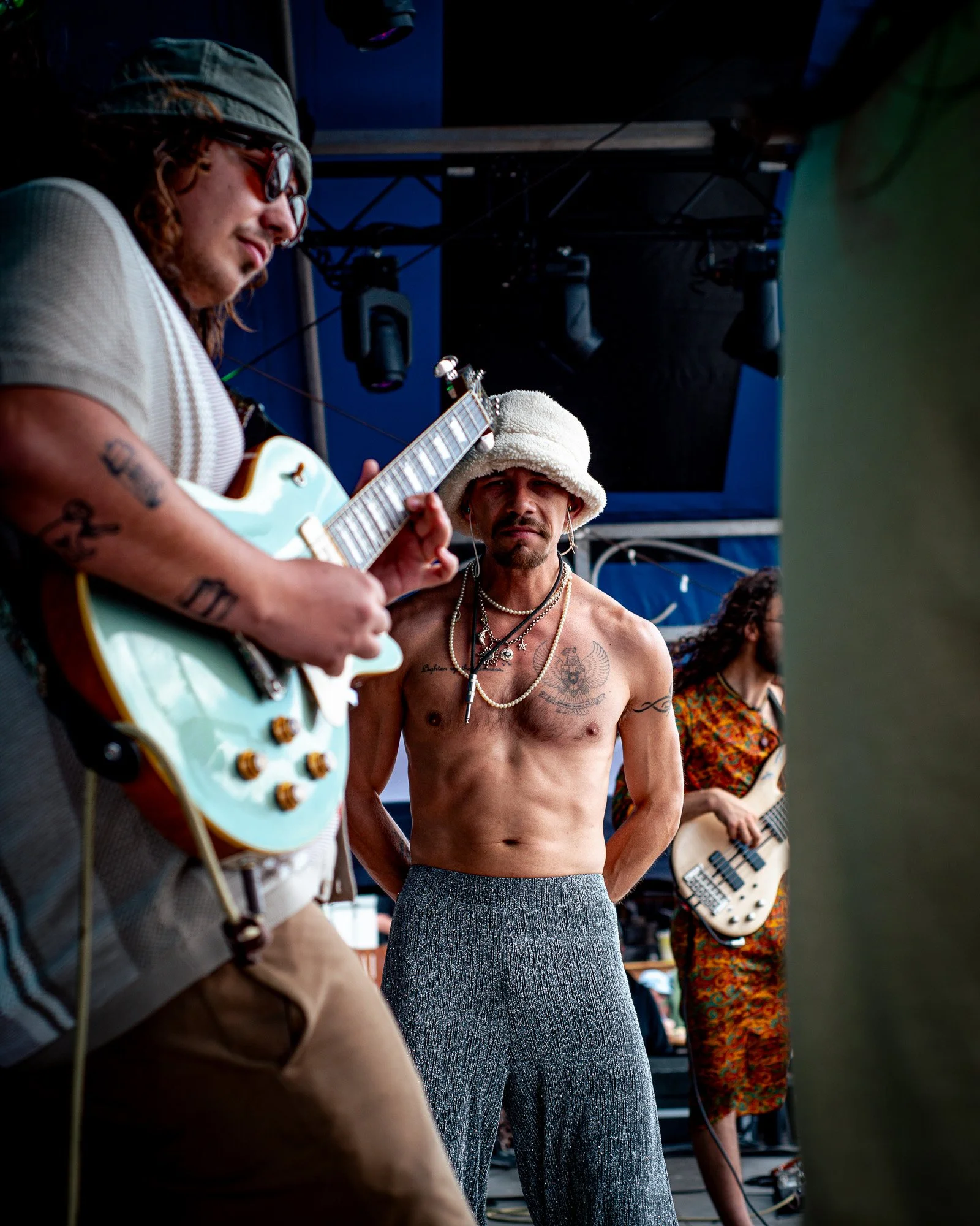 Band Kidd Nebula performing on stage, including a shirtless man with tattoos and jewelry, a man playing bass, and a man playing guitar, with stage lighting and sound equipment visible.