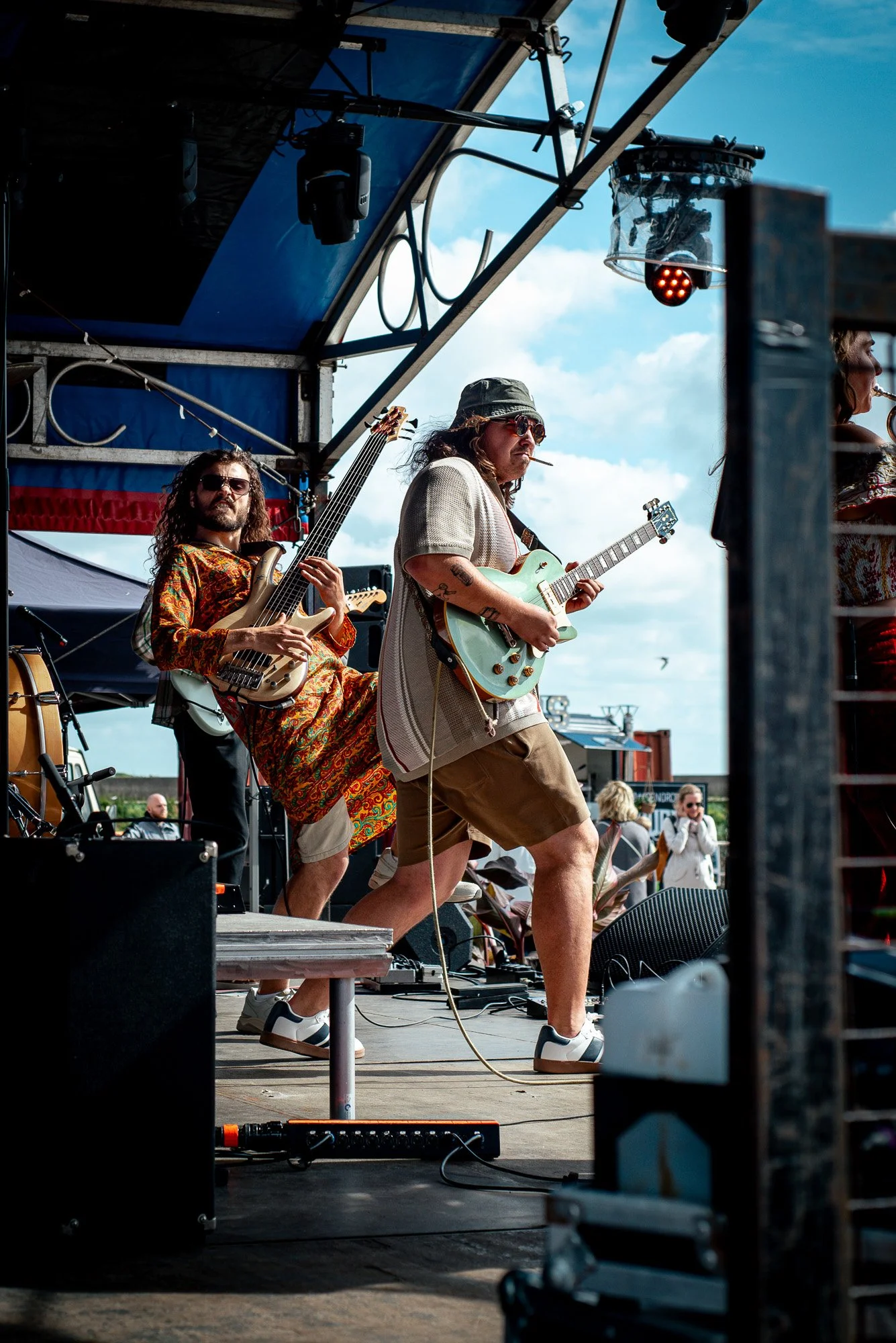 Musicians of the band Toscans Brew performing on outdoor stage under a blue sky, one playing guitar and another playing bass, with an audience in the background.