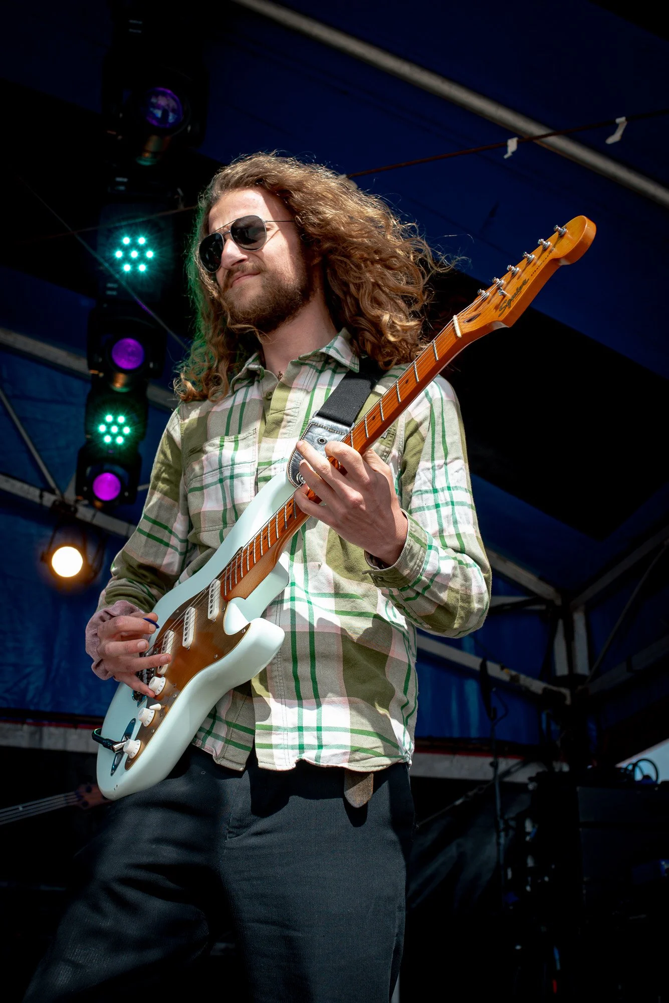 A musician from the band Toscans Brew with long, curly hair and sunglasses playing an electric guitar on a stage at SAIL Harlingen with colorful lights in the background.