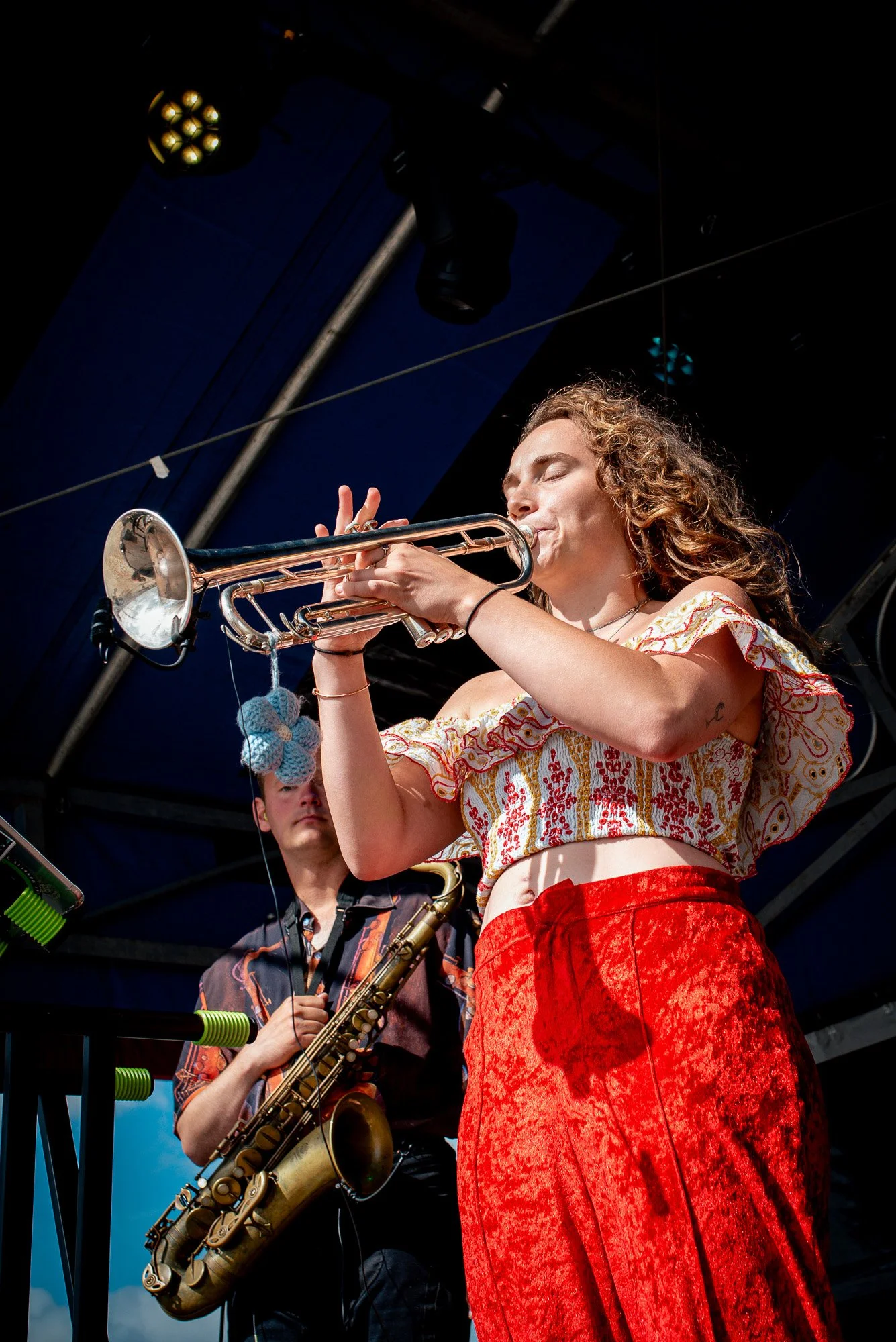 A woman with curly hair playing the trumpet on stage, wearing a colorful off-the-shoulder top and red pants, with a man in the background playing a saxophone. The band is Toscans Brew.