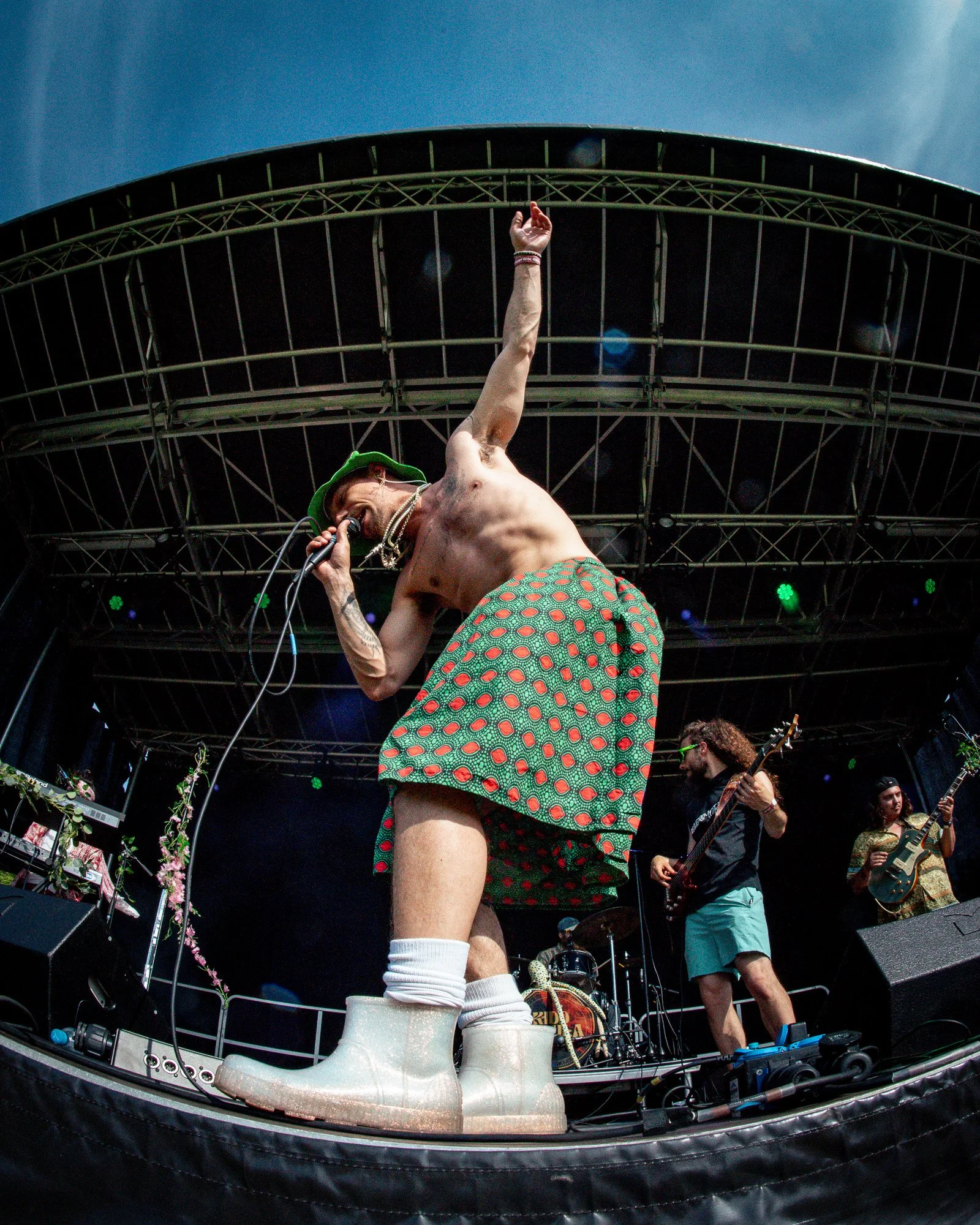 A musician from the band Kidd Nebula performing on stage captured with a fisheye lens, singing into a microphone, with band members playing guitars in the background, and athletic shoes placed at the front of the stage.