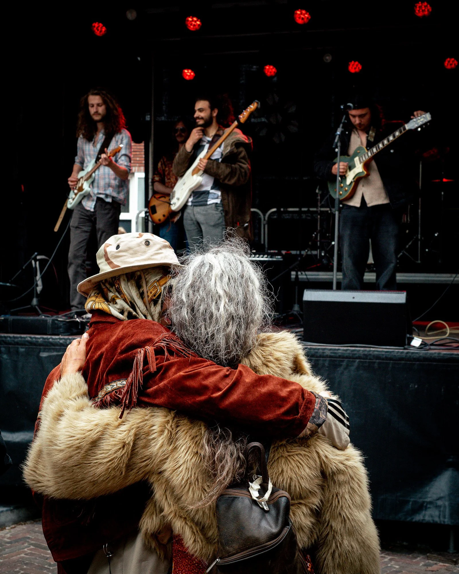 A person with long gray hair and a fur coat hugging another person with a hat in front of a stage with musicians playing guitars and singing, red stage lights overhead.