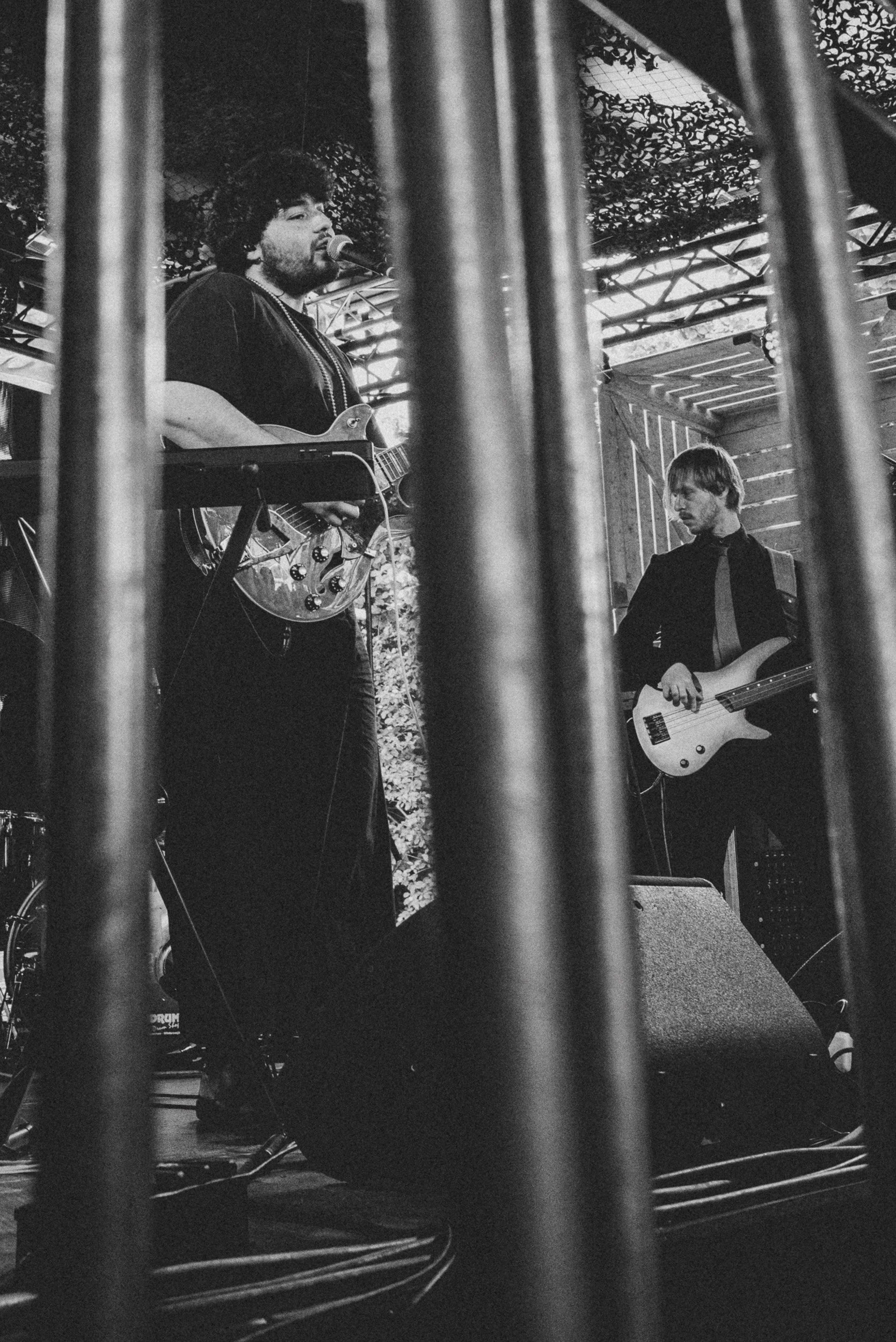 Black and white photo of two musicians from the band Zerguzest performing on stage, one playing guitar and singing into a microphone, the other playing a fretless bass guitar, viewed through vertical bars.