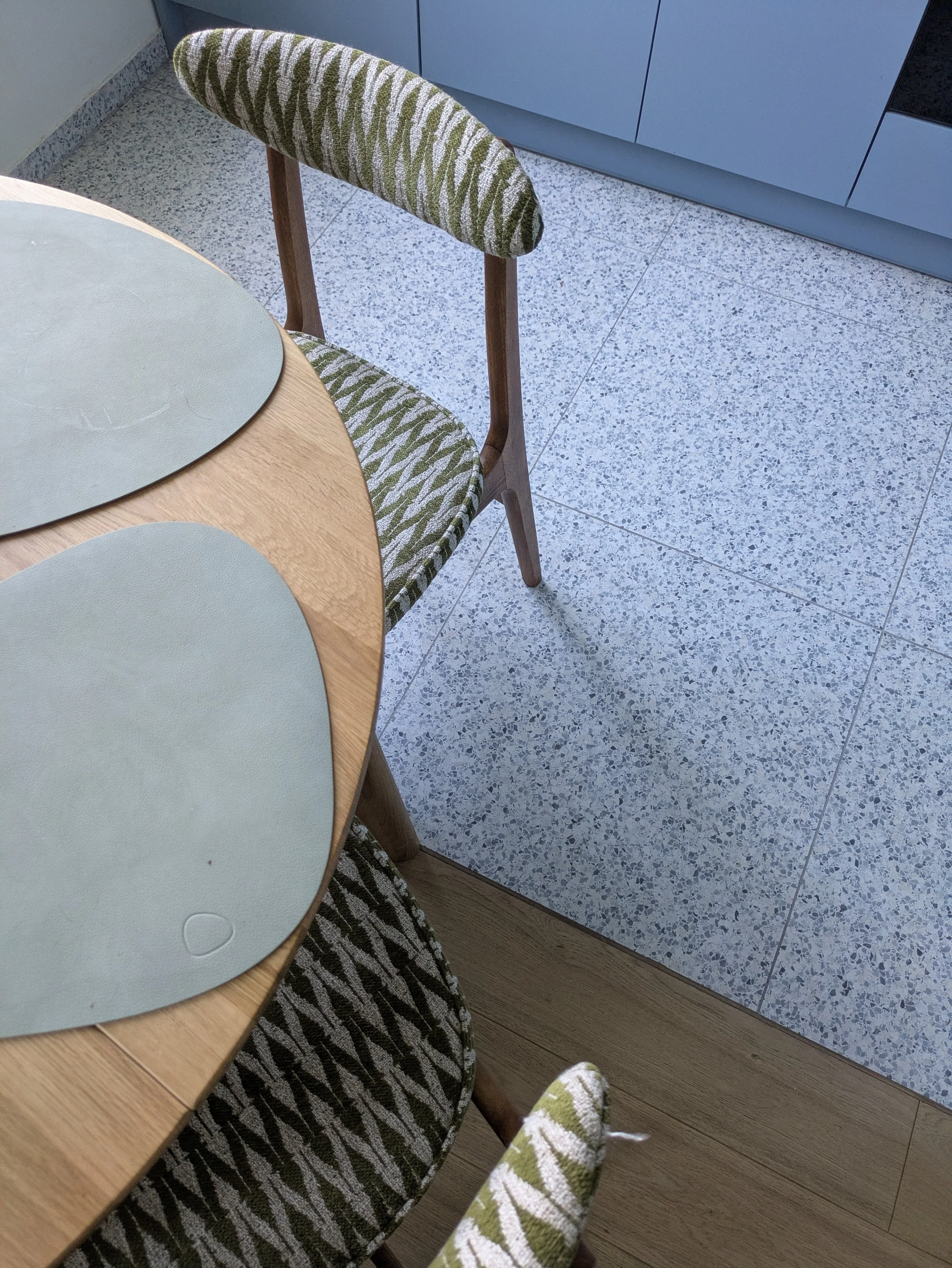 A kitchen and dining area with a wooden table, two light blue placemats, and a chair with a patterned green, white, and brown fabric cushion. The flooring is speckled gray tile, and there are blue cabinets in the background.
