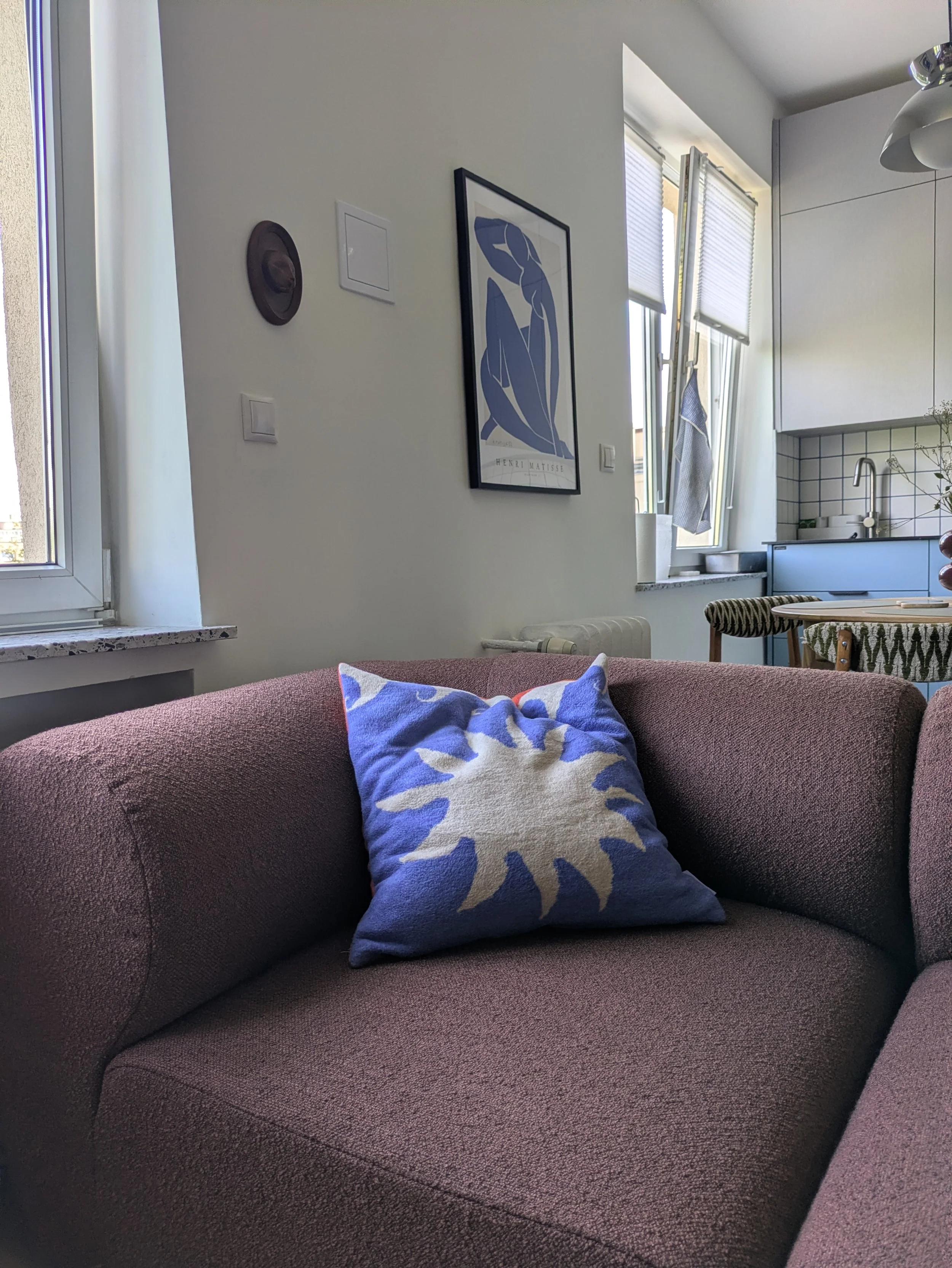 Living room with a pink fabric sofa, a decorative pillow with a white and blue design, a wall with a Henri Matisse art print, a window with open blinds, and a kitchen area with white cabinets and a sink.