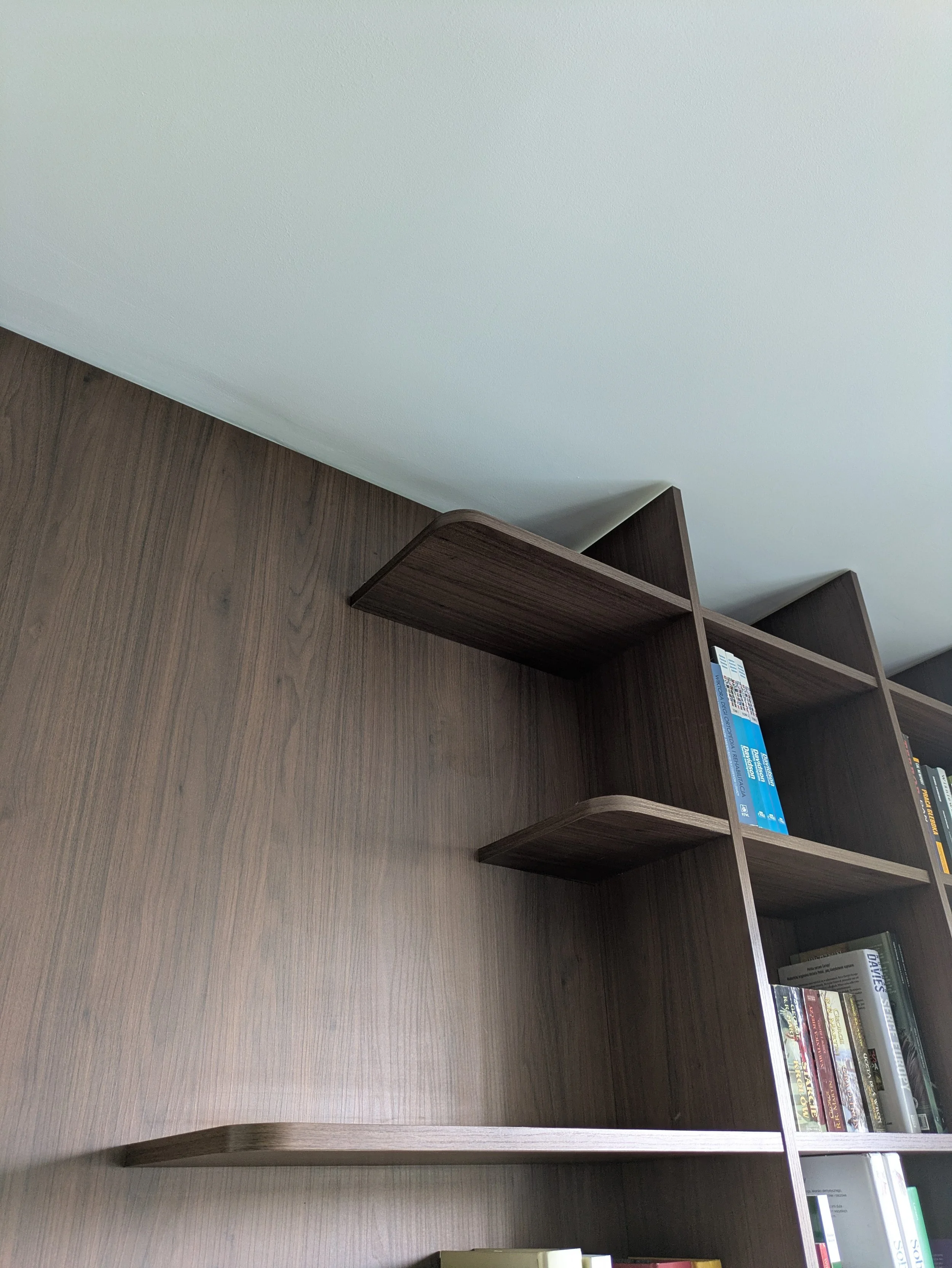 A wooden bookshelf with several books on the right side and a plain wall on the left. The bookshelf is empty on the left side and has books on the right.