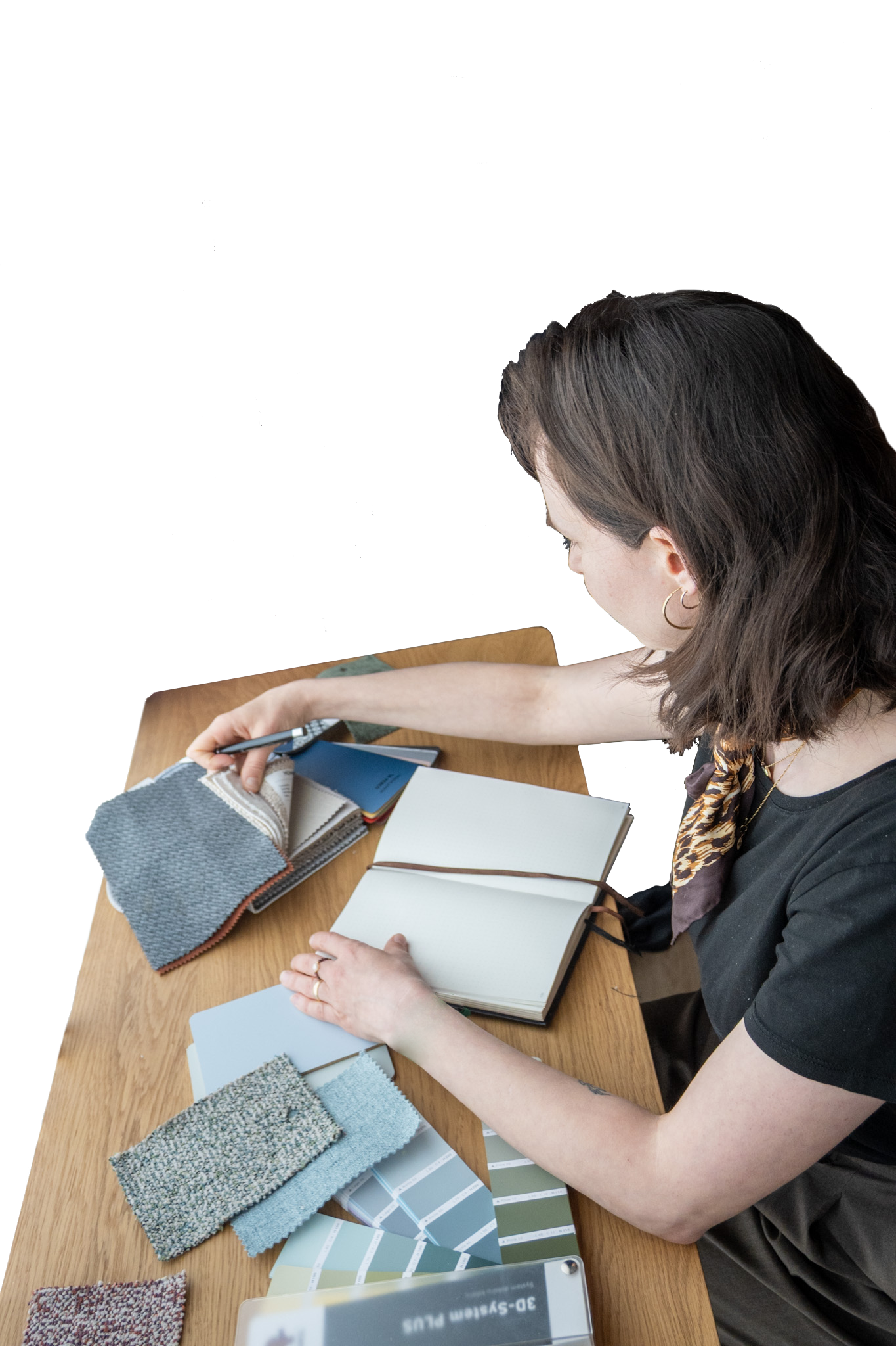 A woman with shoulder-length brown hair and wearing a black top with animal print scarf is seated at a wooden table. She is examining fabric swatches, color palettes, and paint samples. The table has fabric samples, paint chips, a notebook, and a phone.