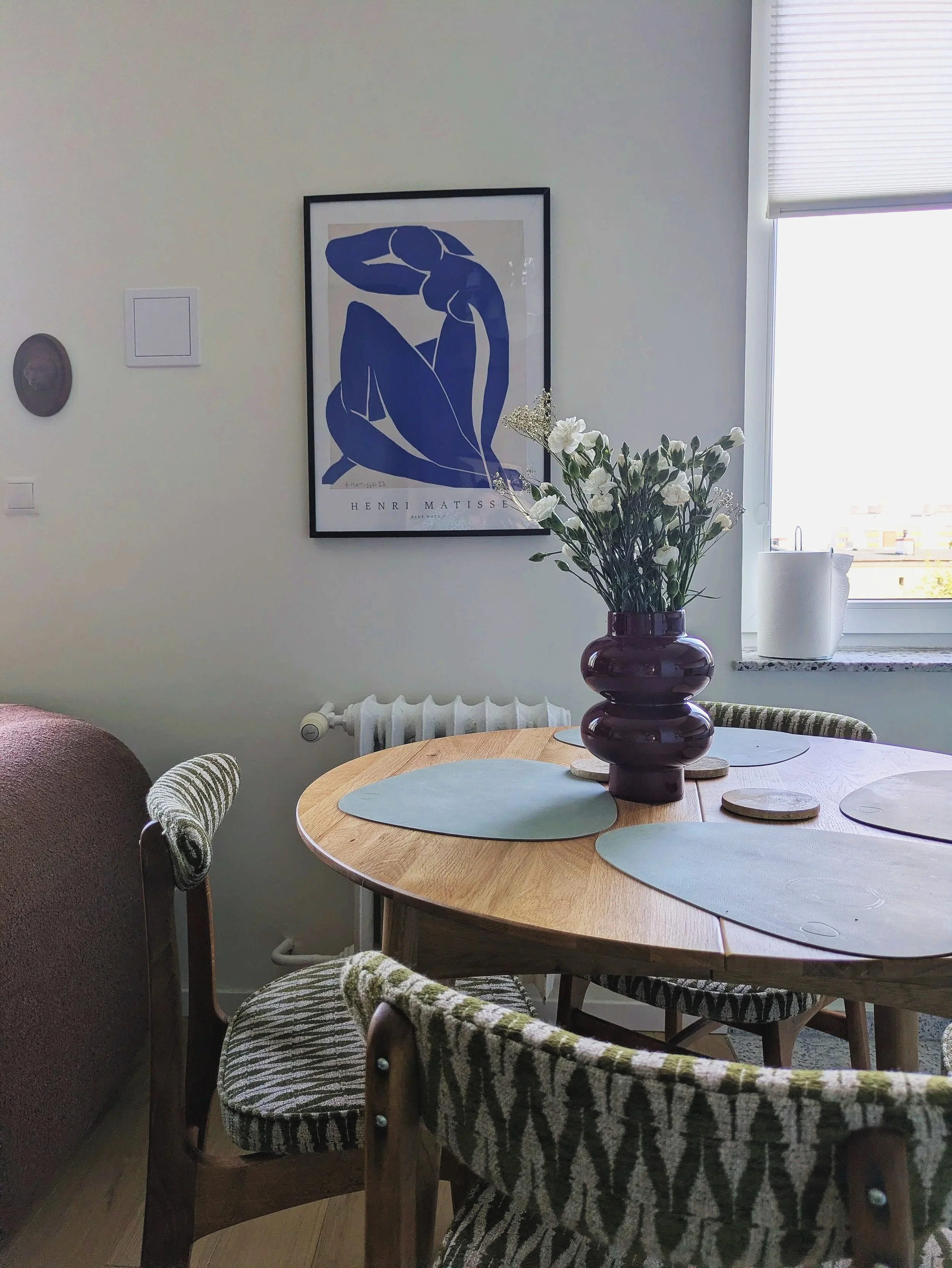 A dining area with a round wooden table and patterned chairs. A large dark purple vase with white flowers is on the table. In the background, there's a framed Henri Matisse artwork, a window with blinds, and a radiator underneath.