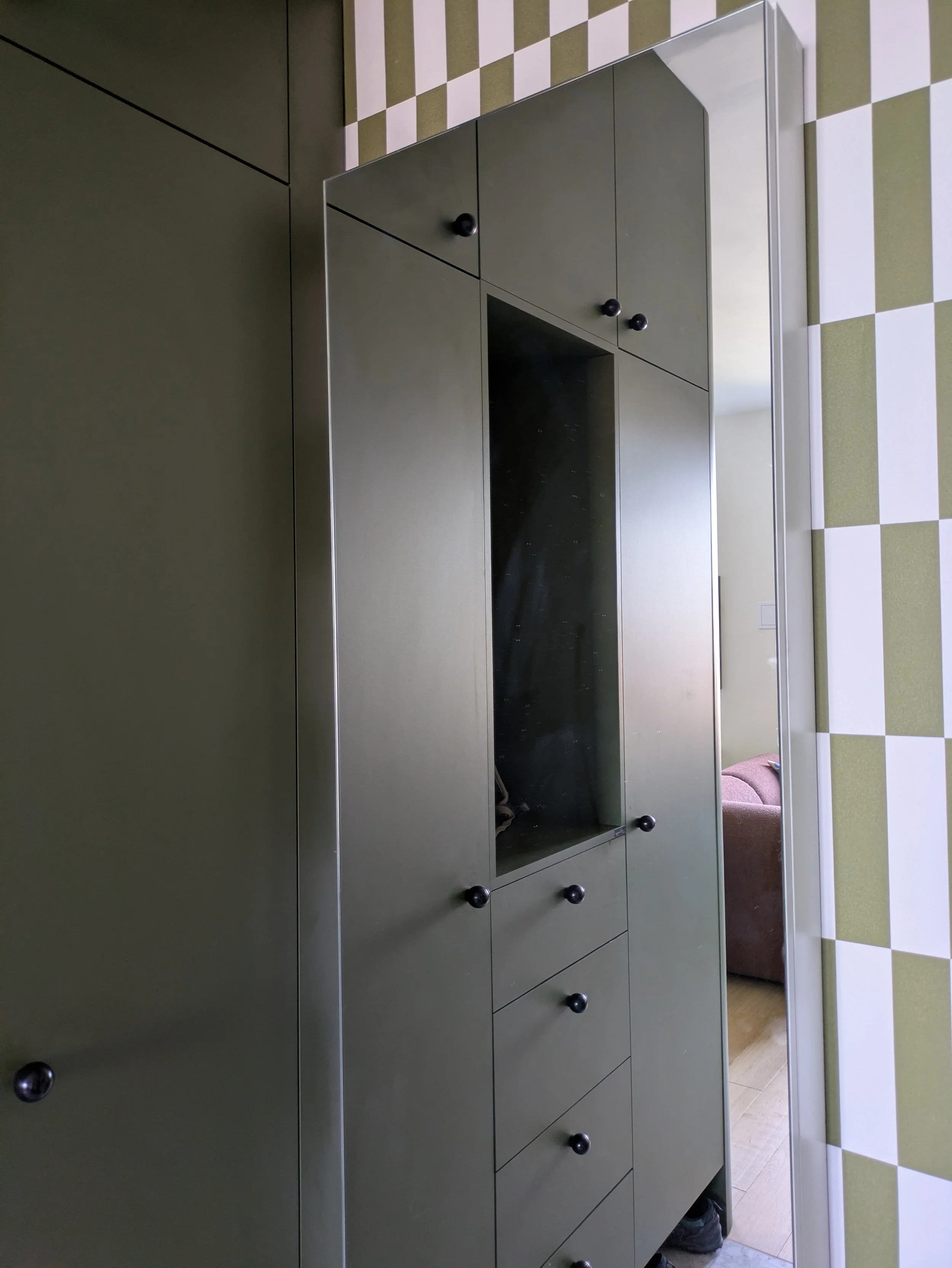 A green cabinet with black knobs and a mirror, situated in a room with green and white checkered floor tiles. Part of a pink sofa is visible through the door opening.