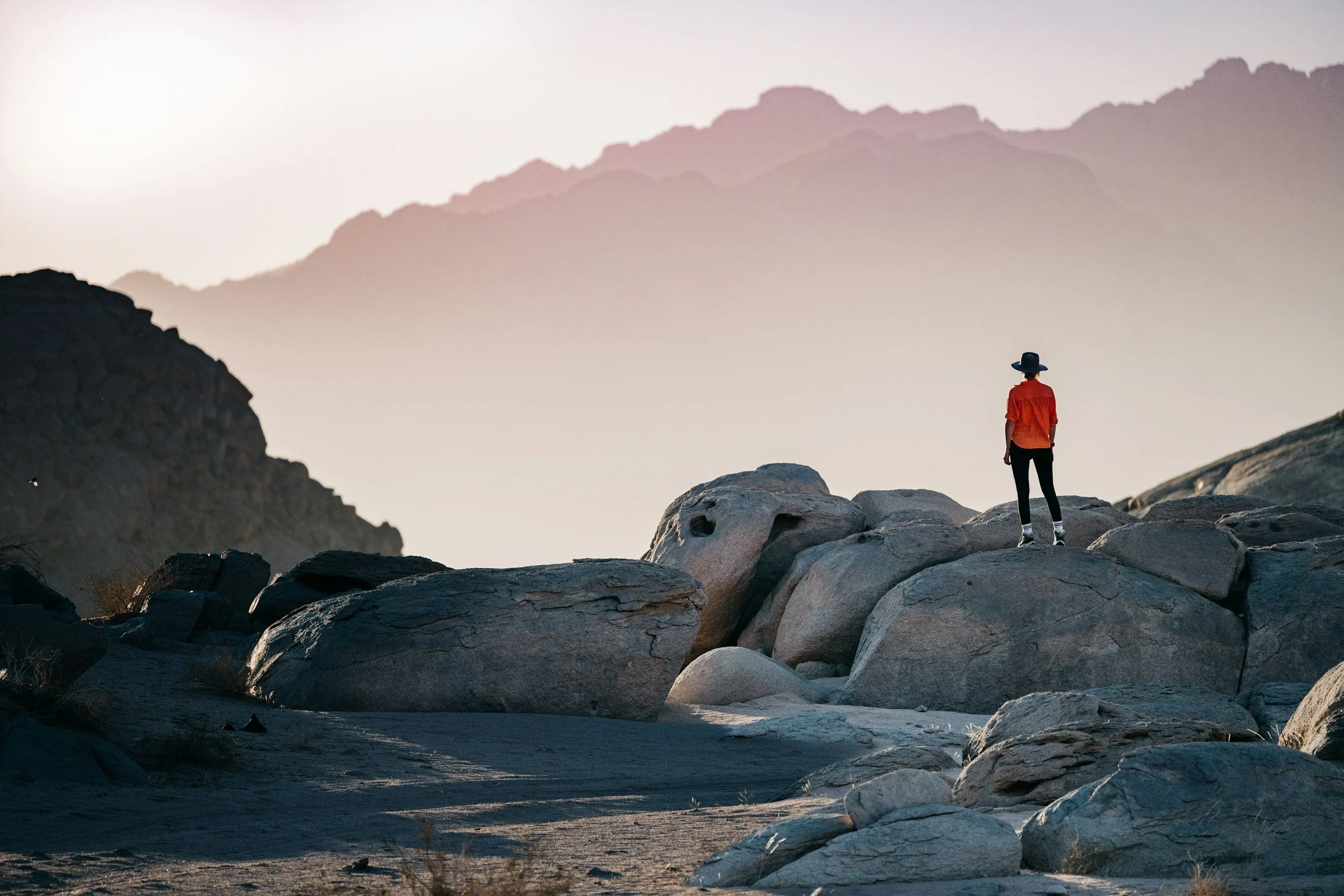 Person wearing a hat and orange jacket standing on rocks in a mountainous landscape during dusk or dawn