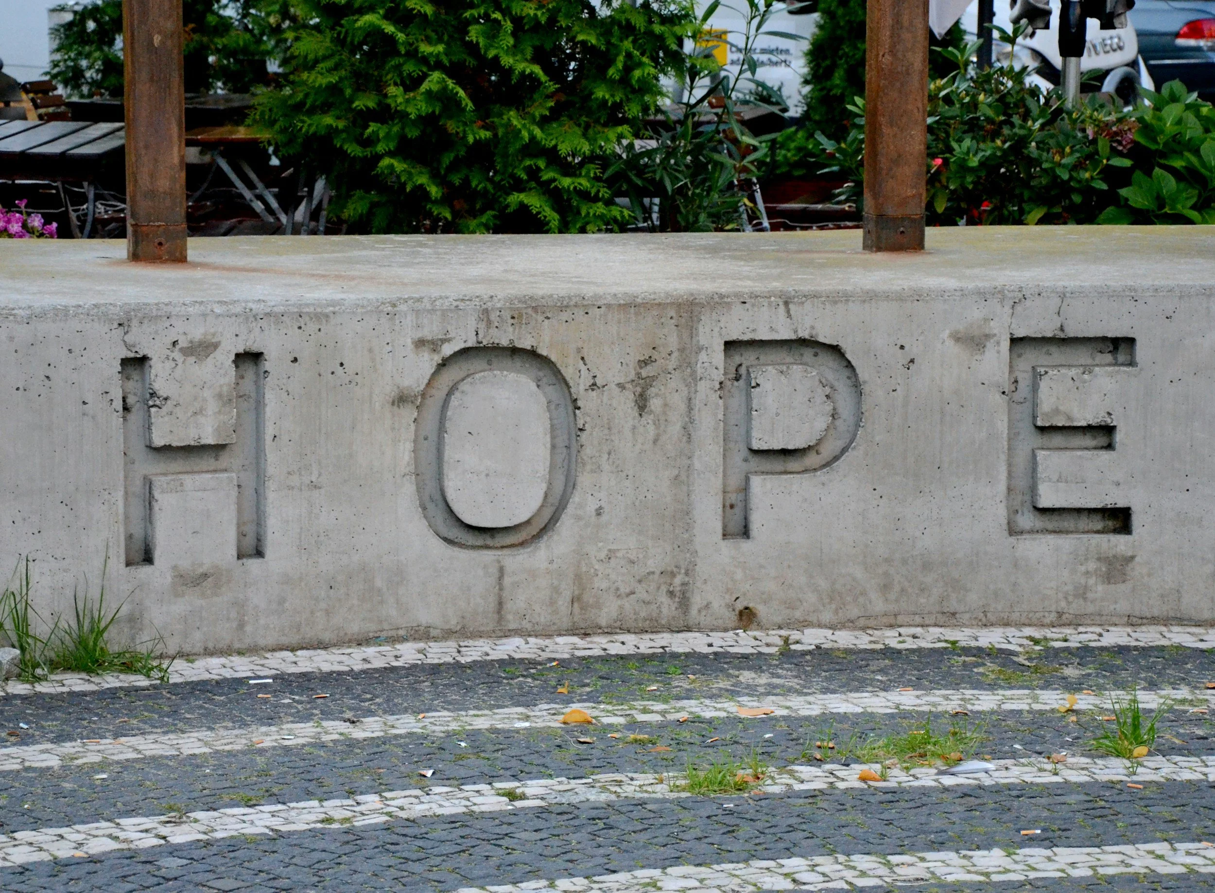 A concrete barrier with the partial engraved word 'HOPE' at the bottom, a sidewalk with small cobblestones and grass at the edge, behind which there are green bushes, trees, and parked vehicles.