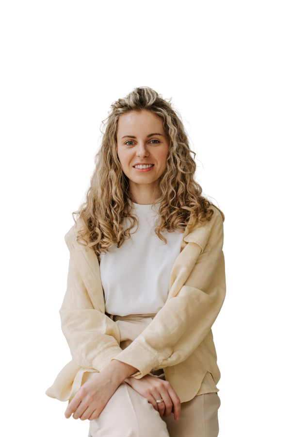 A woman with curly blonde hair, wearing a white shirt and beige jacket, sitting with her legs crossed and smiling at the camera.