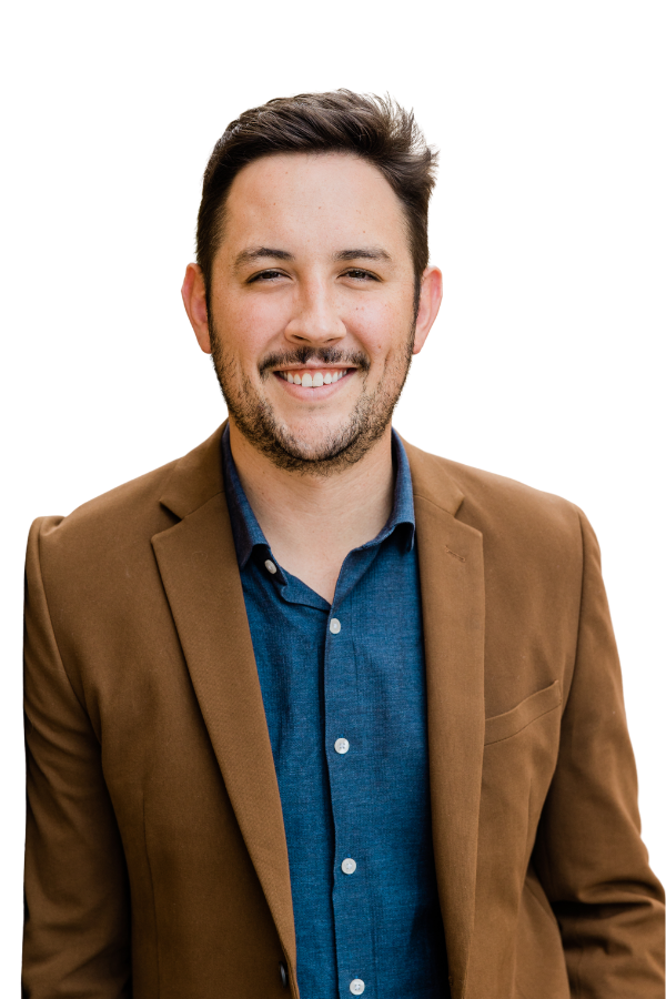 Smiling man with brown hair, beard, wearing a brown blazer and blue shirt, standing against a plain background.