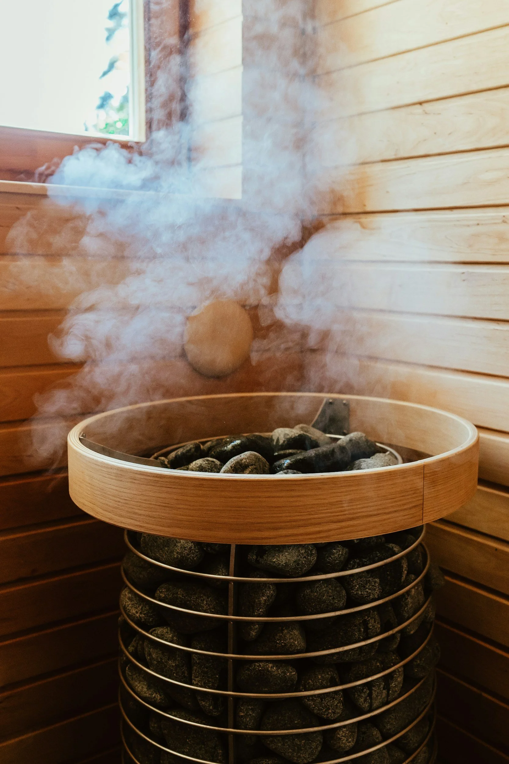 Steam rising from a sauna heater filled with black stones inside a wooden sauna room.