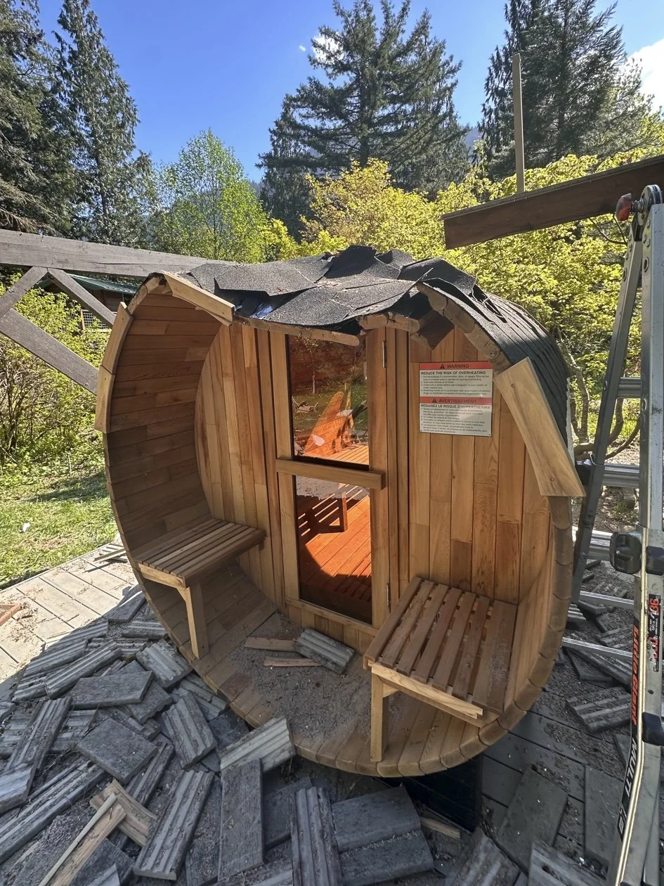 A small, cylindrical wooden sauna under construction, with an arched roof and two benches inside, situated outdoors among trees.