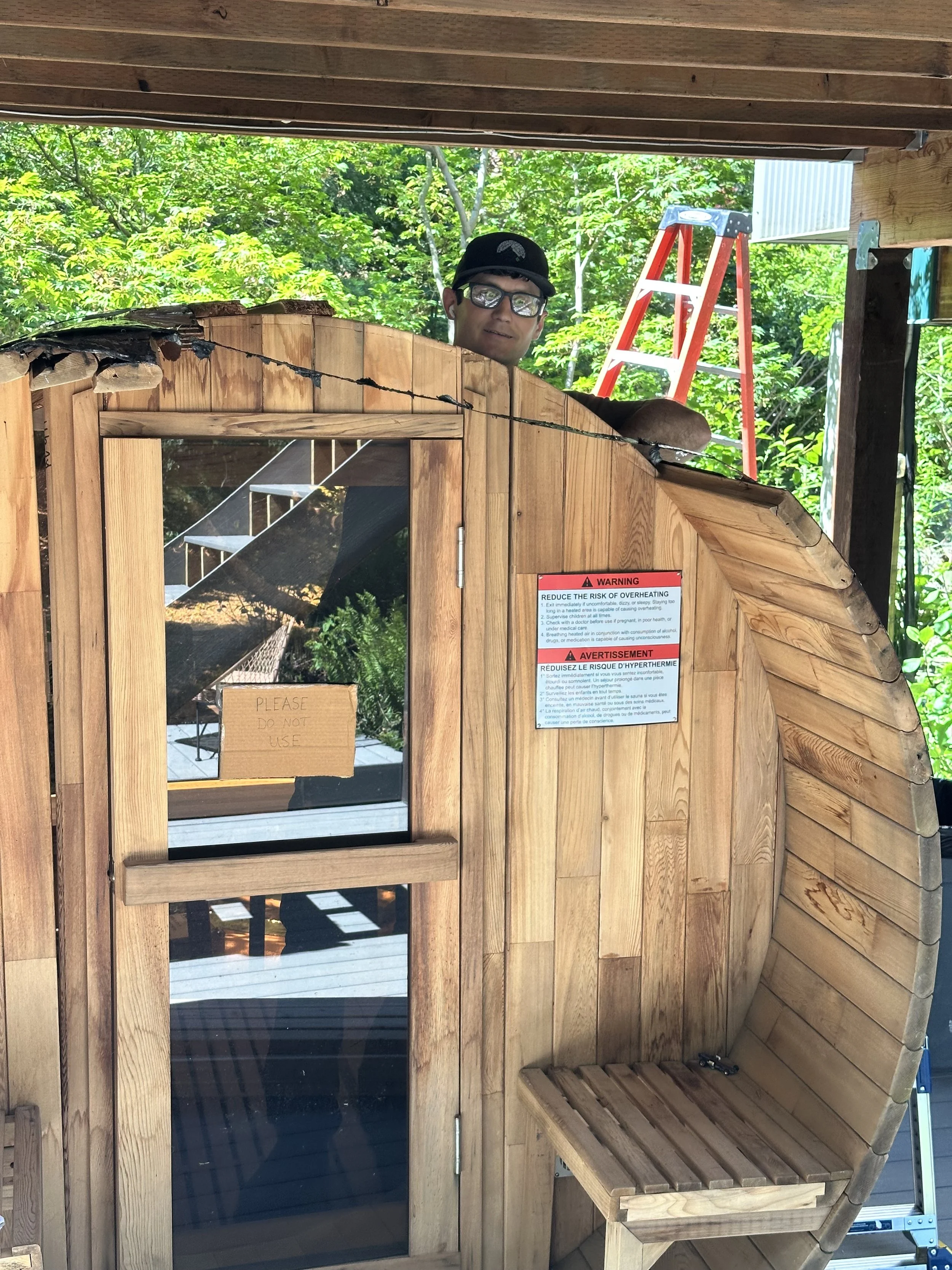 A small wooden outdoor shelter with a sloped roof, a window, and a partial bench. There is a sign that says 'Please Do Not Use' on the door and a warning label about overheating.